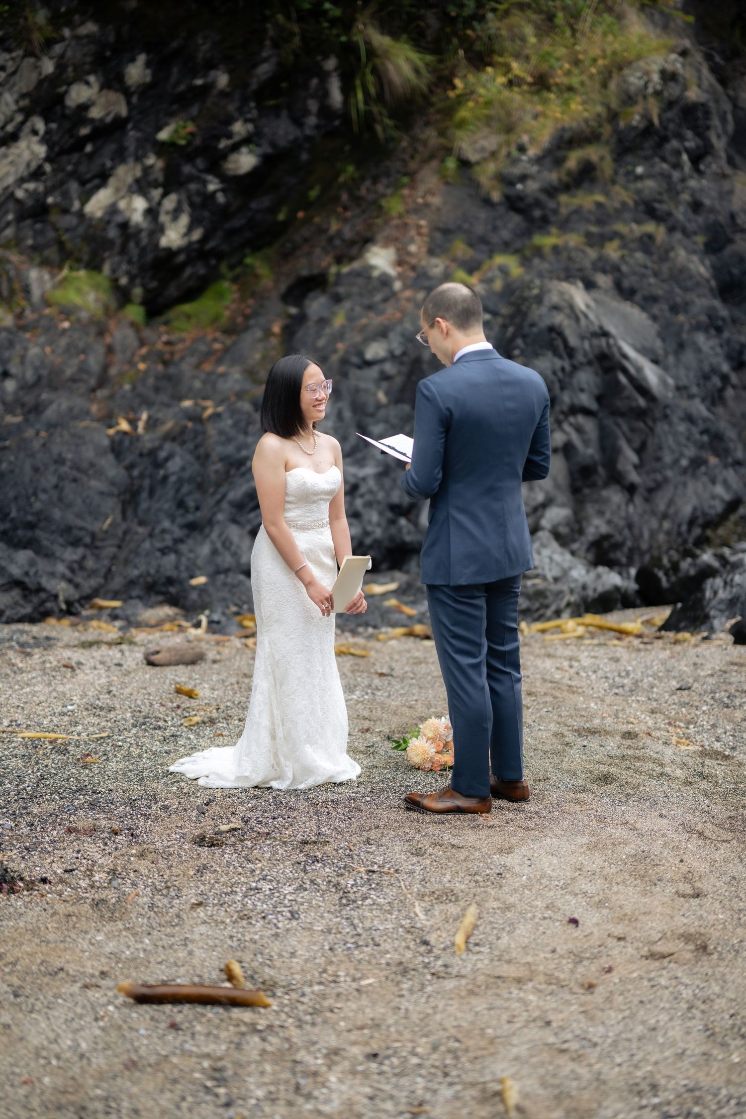Couple stands together during their intimate beach wedding ceremony against rocky coastal backdrop.