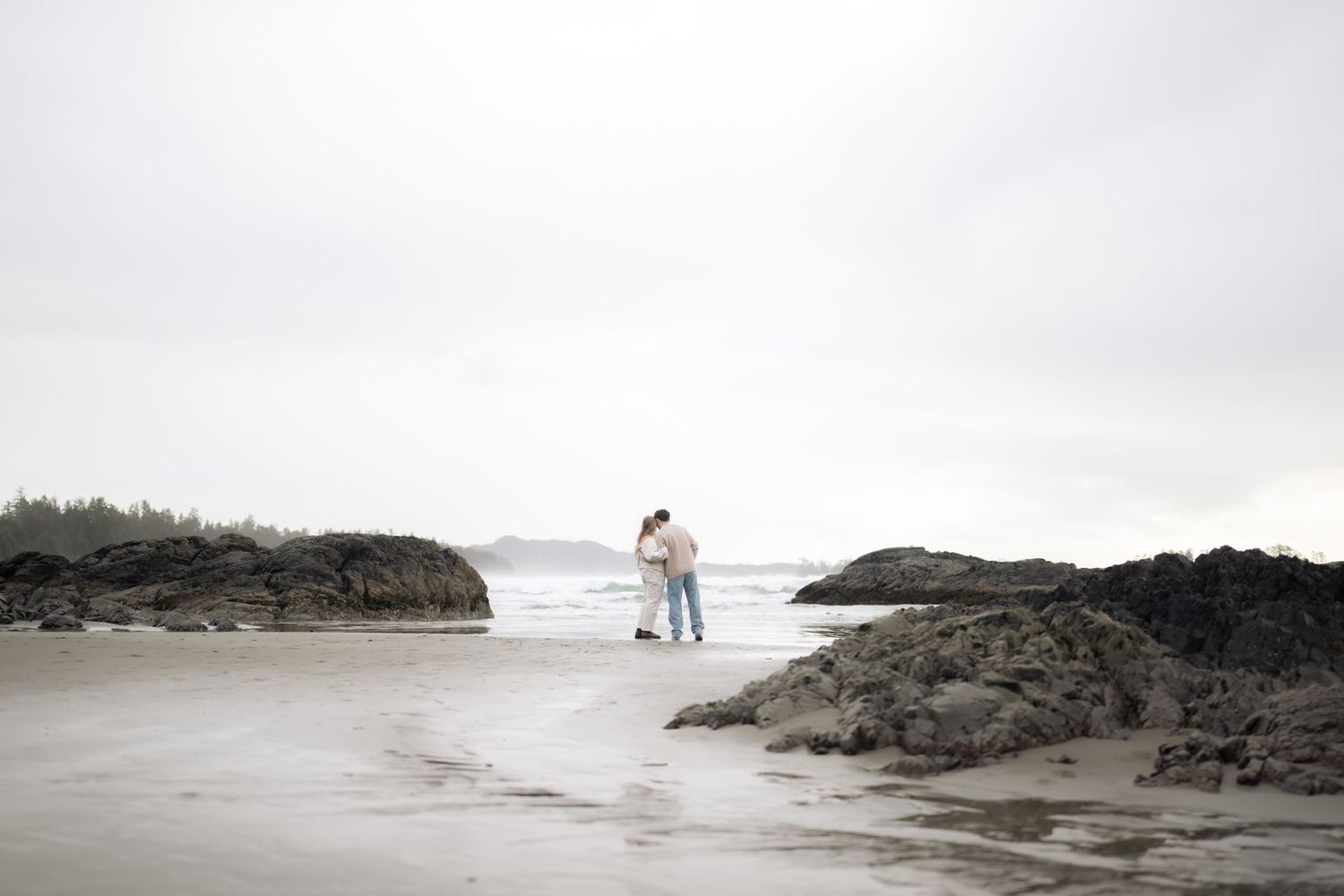 Romantic beach scene with figures standing near rocky outcrops on a foggy day.