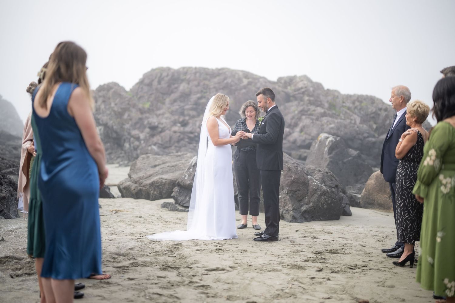 Intimate beach wedding ceremony with attendees gathered near rocky coastal formations.