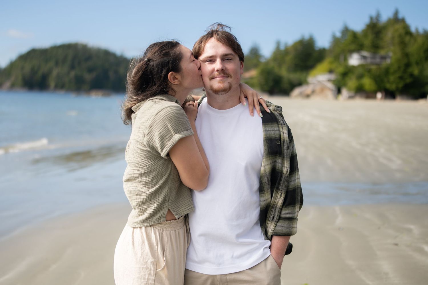 A couple embraces on a scenic beach with mountains and forest in the background.