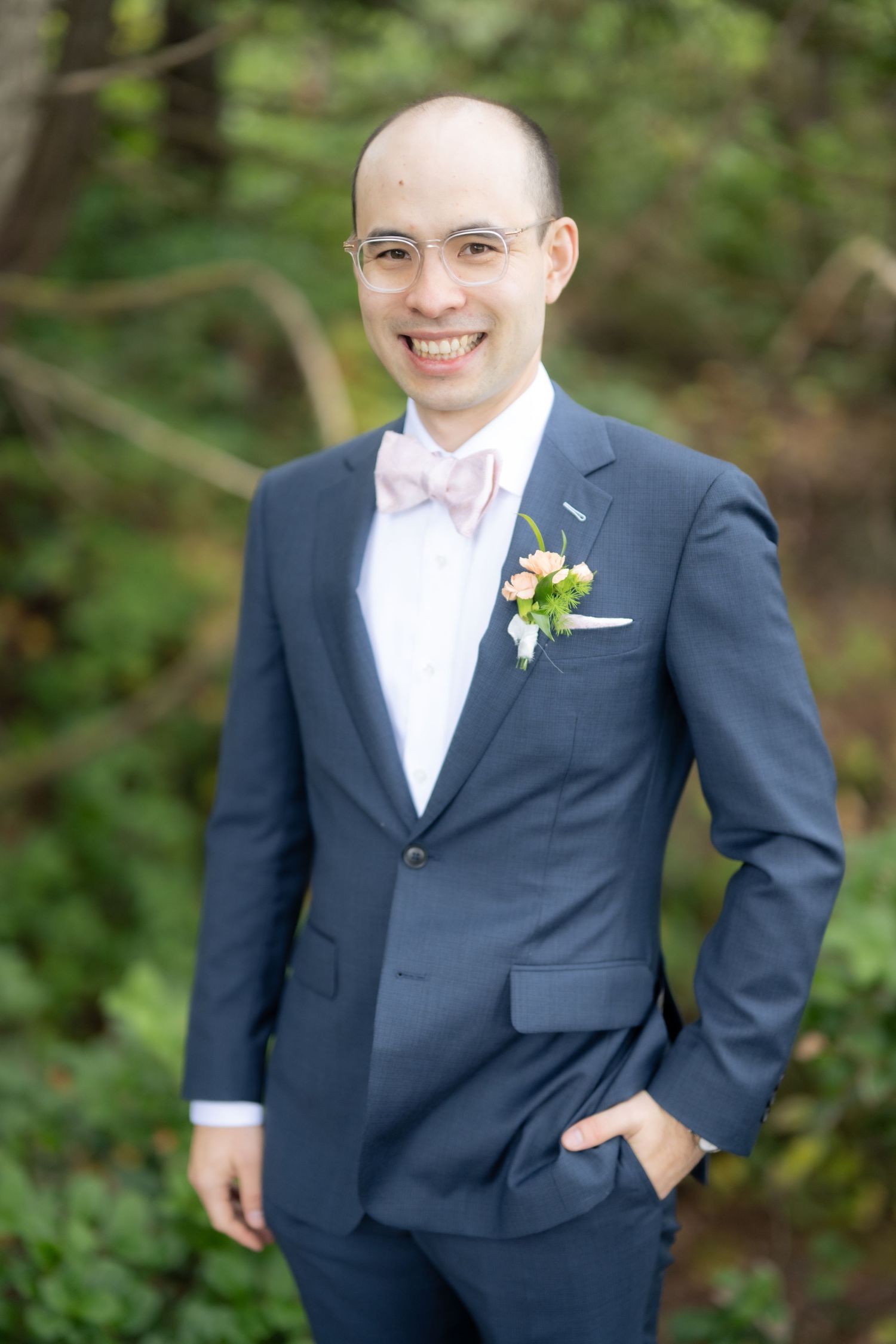A person in a navy suit and white bowtie smiles against a green forest background.