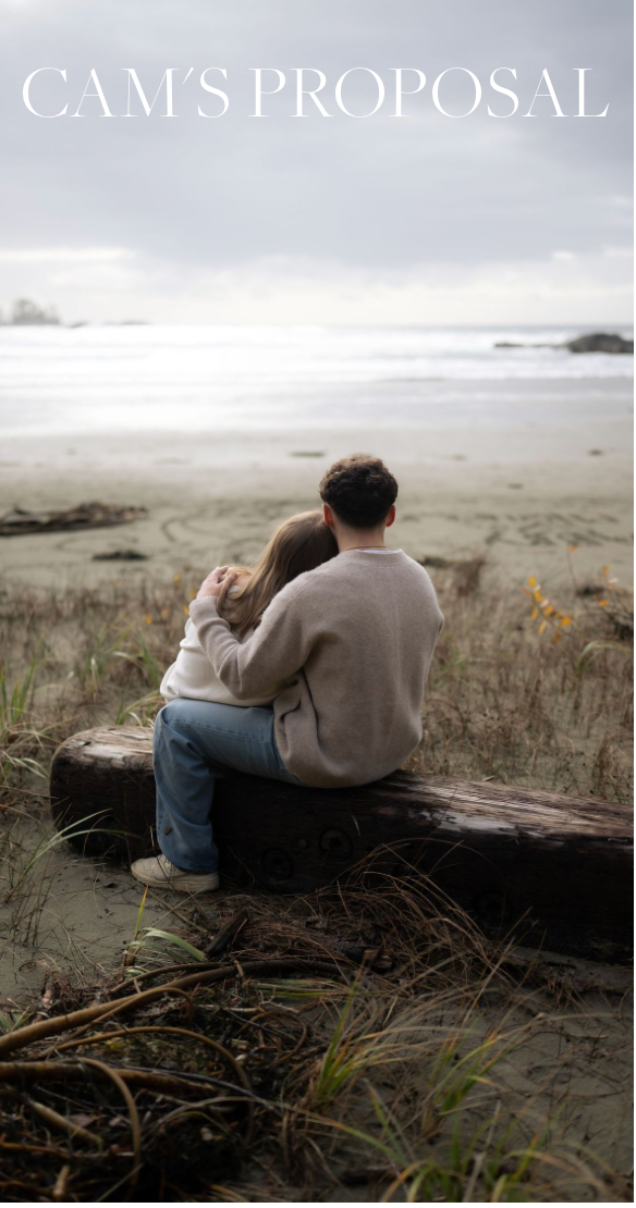 A couple sits close together on a log at a misty beach during what appears to be a romantic marriage proposal moment.