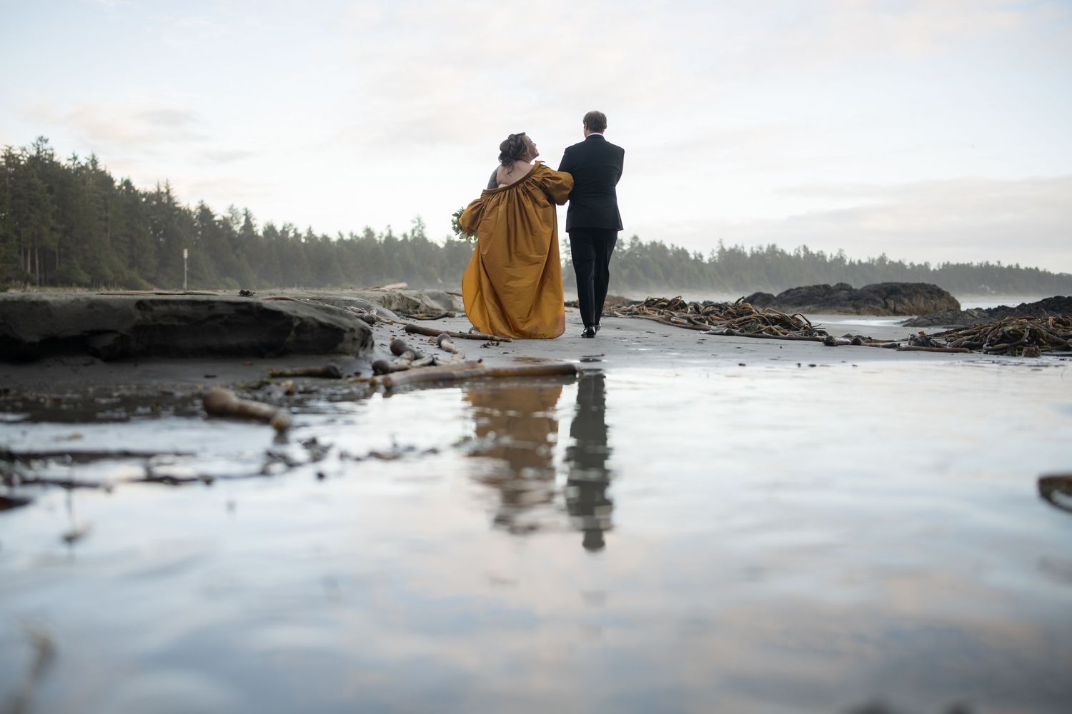 Two silhouettes walk along a misty beach with evergreen trees in the background.