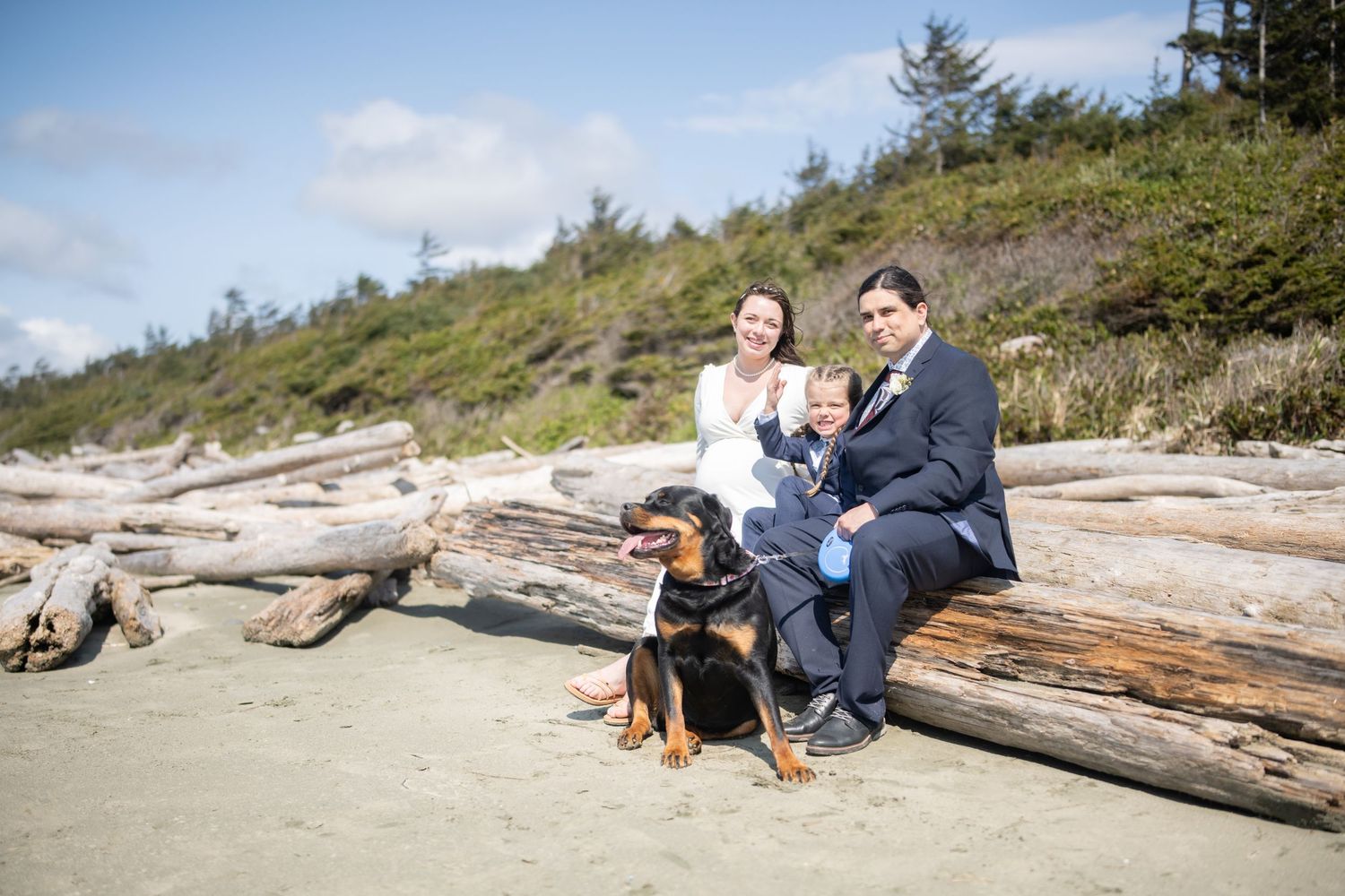Group gathered near driftwood logs on beach with forested hillside in background.
