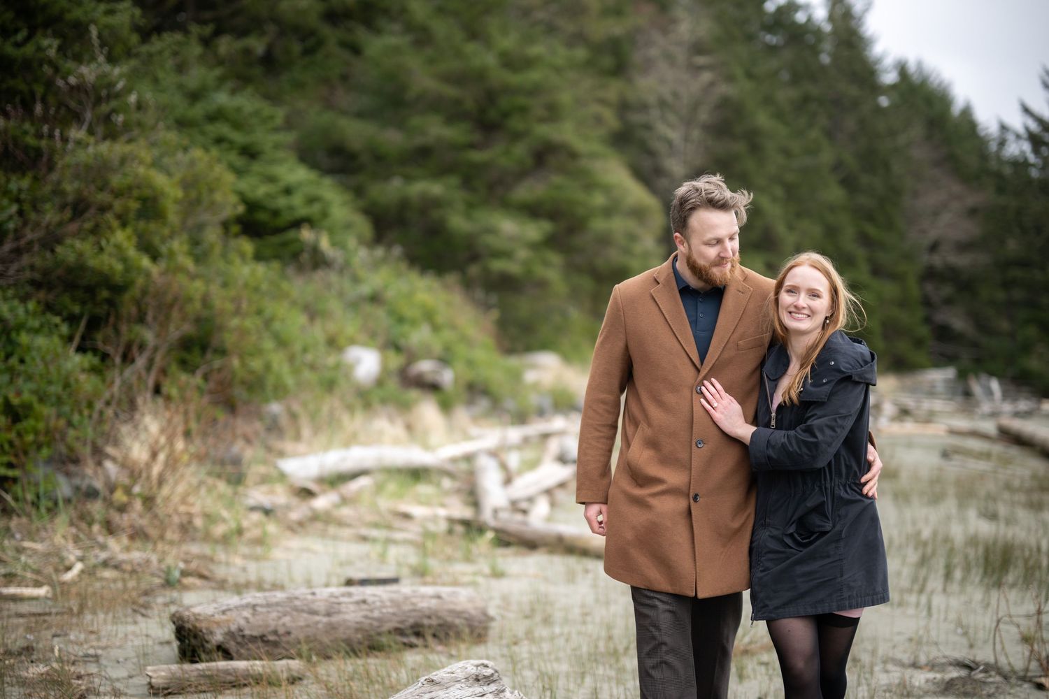 A couple walks together along a rocky coastal path surrounded by evergreen trees.