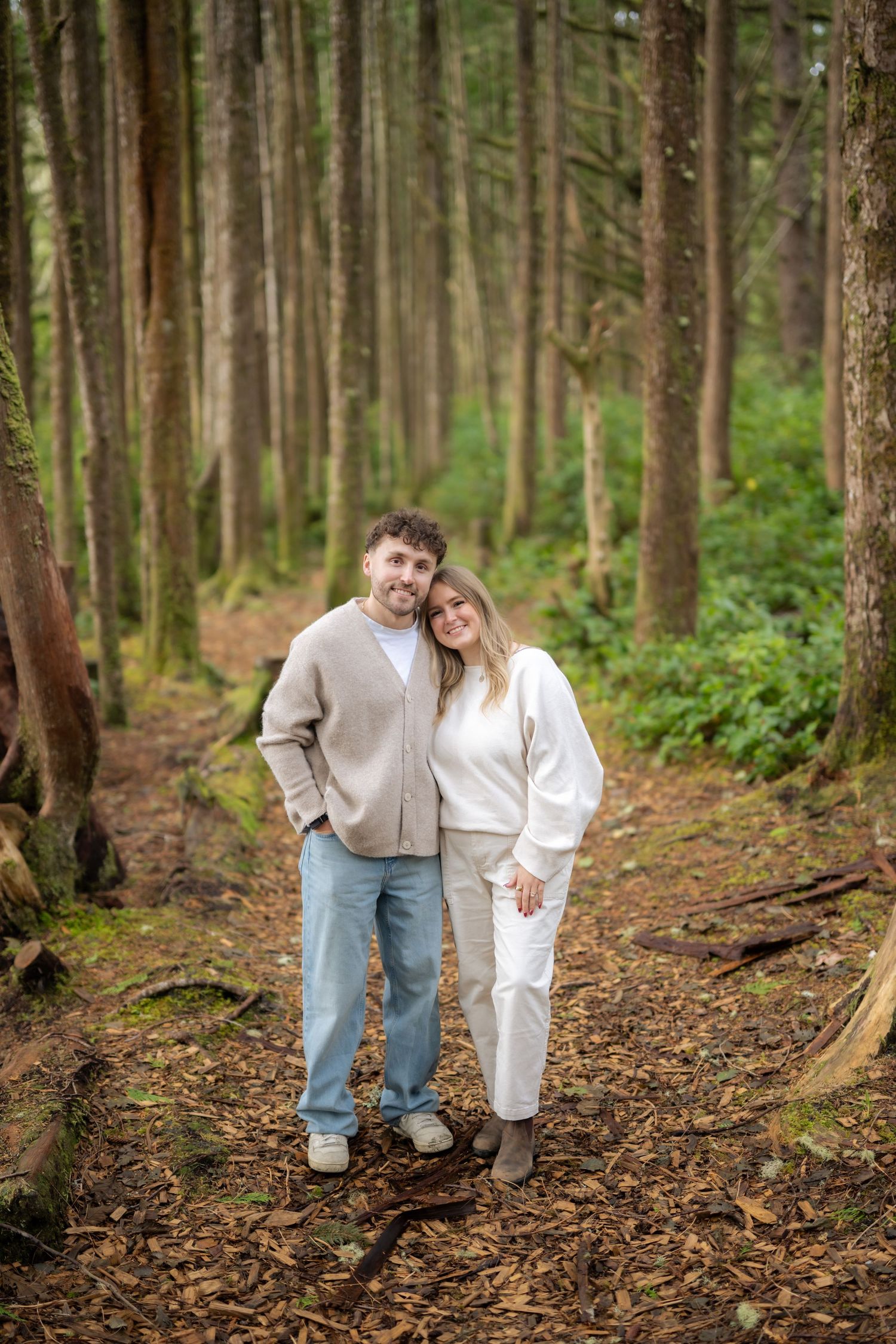 Couple walking together through pine forest wearing matching light colored outfits.