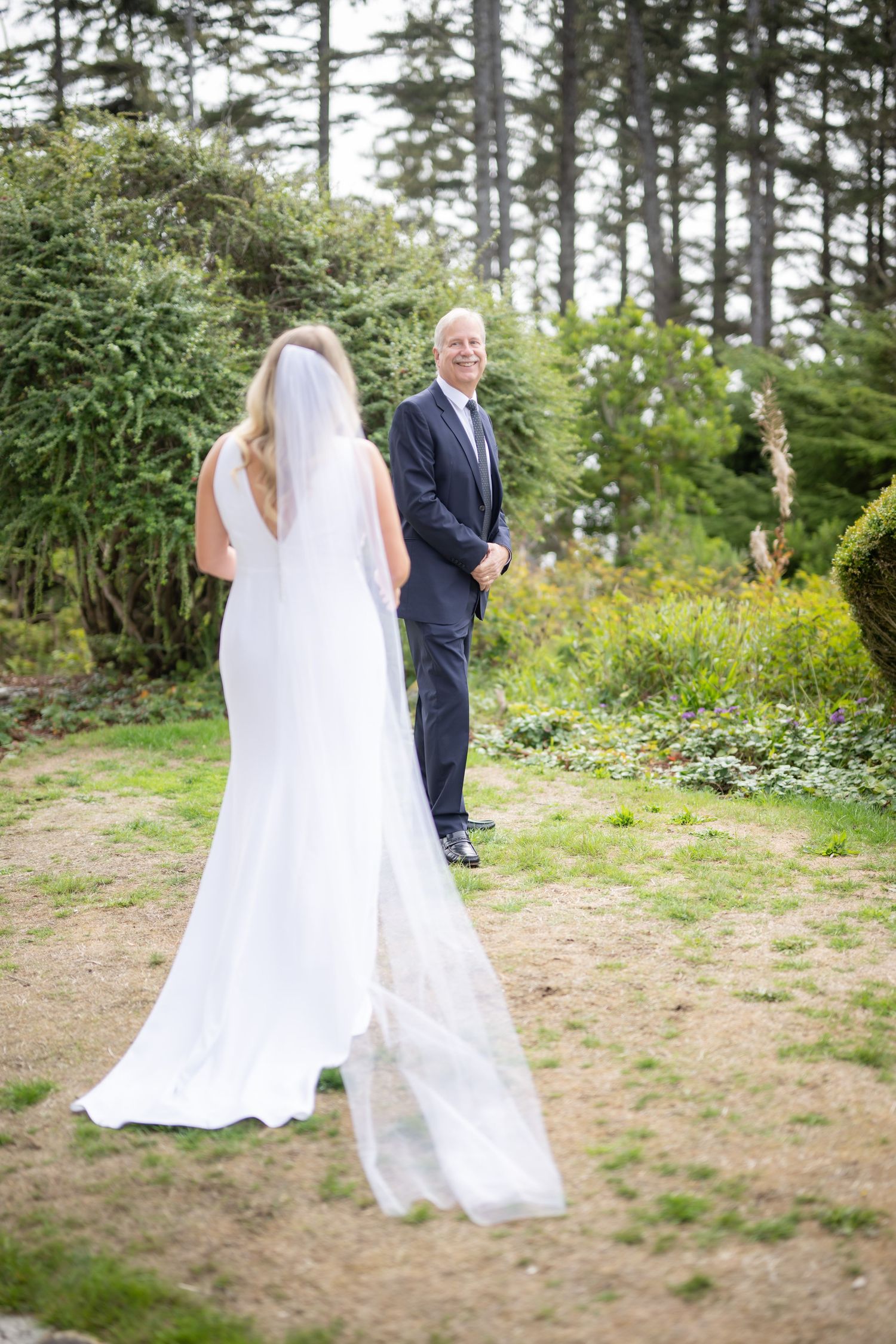 Bride in flowing white dress approaches groom for first look in forest setting.