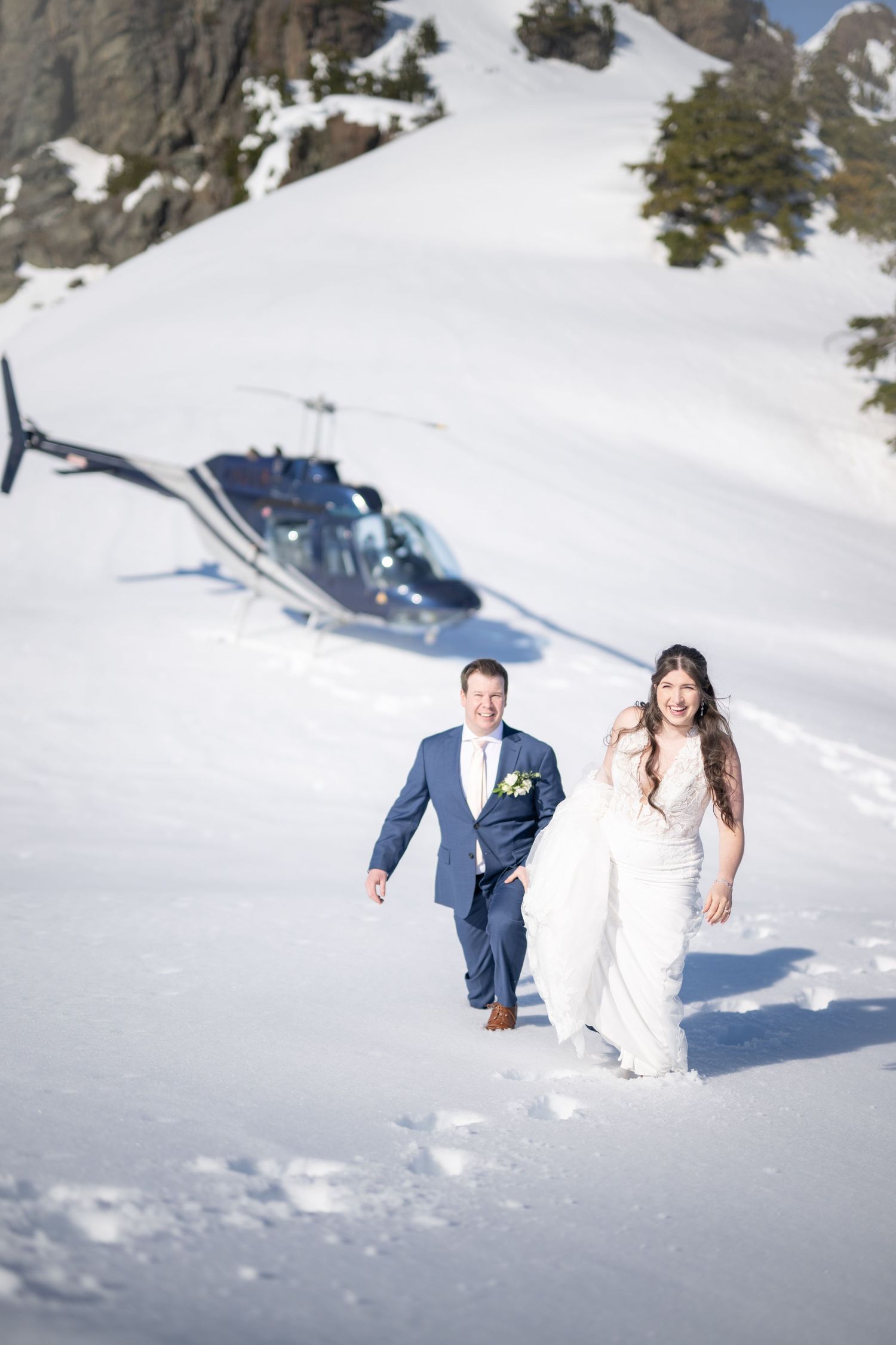 Playful wedding couple posing in deep snow with mountains and evergreen trees in the background.