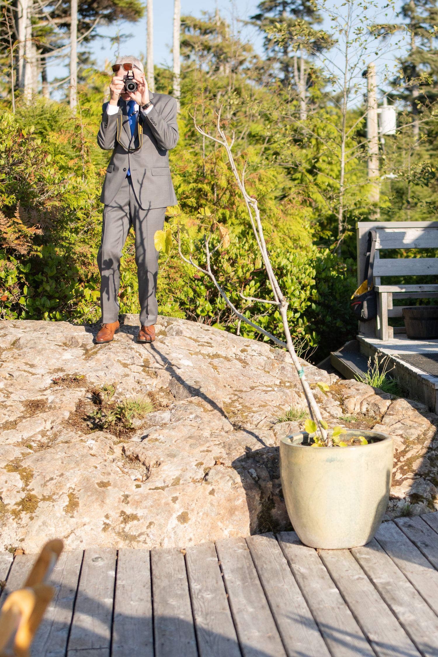 Photographer in grey suit takes pictures on wooden deck with potted plant.