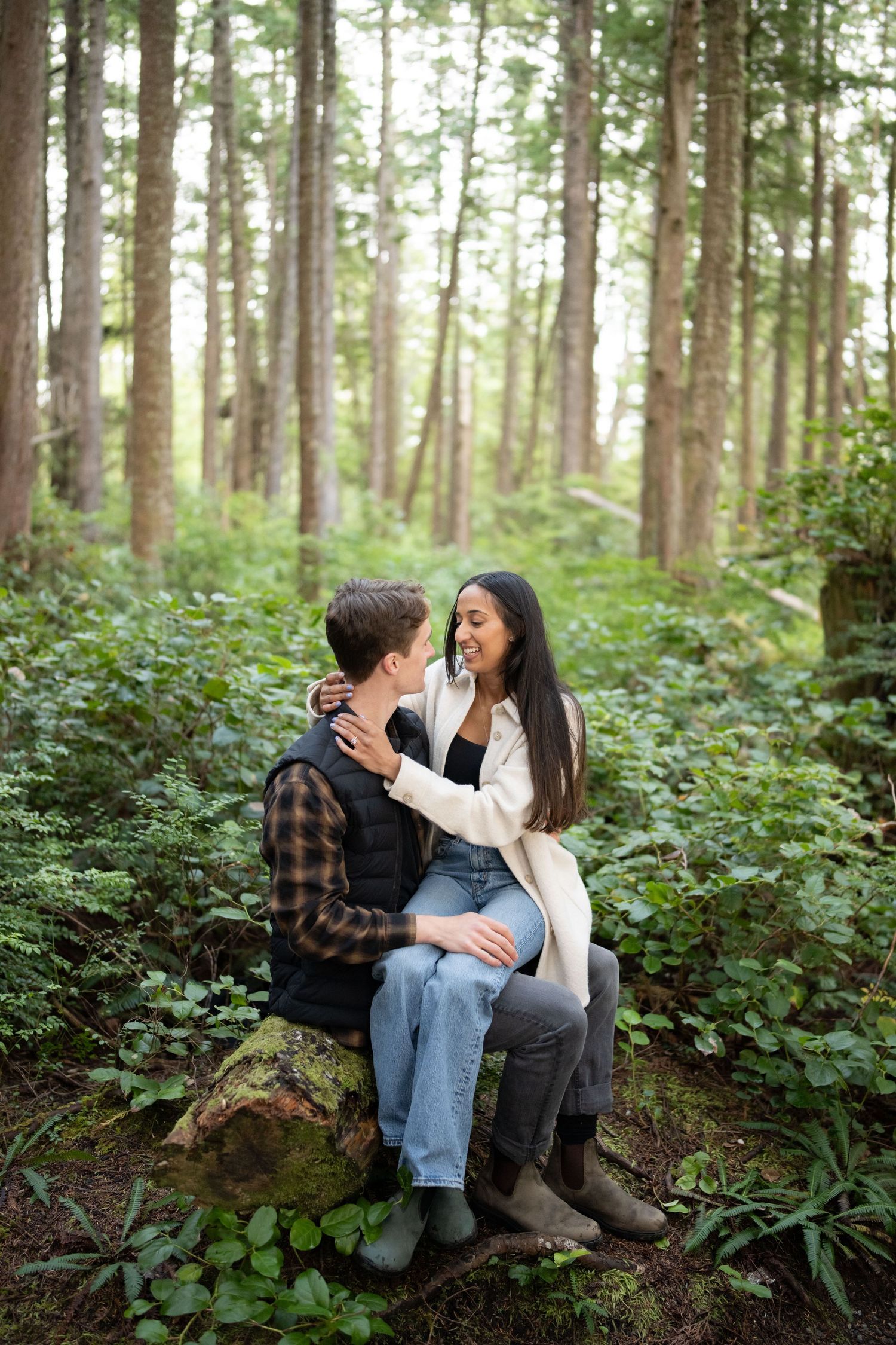 Two people laughing together while seated on a mossy log in a dense forest grove.