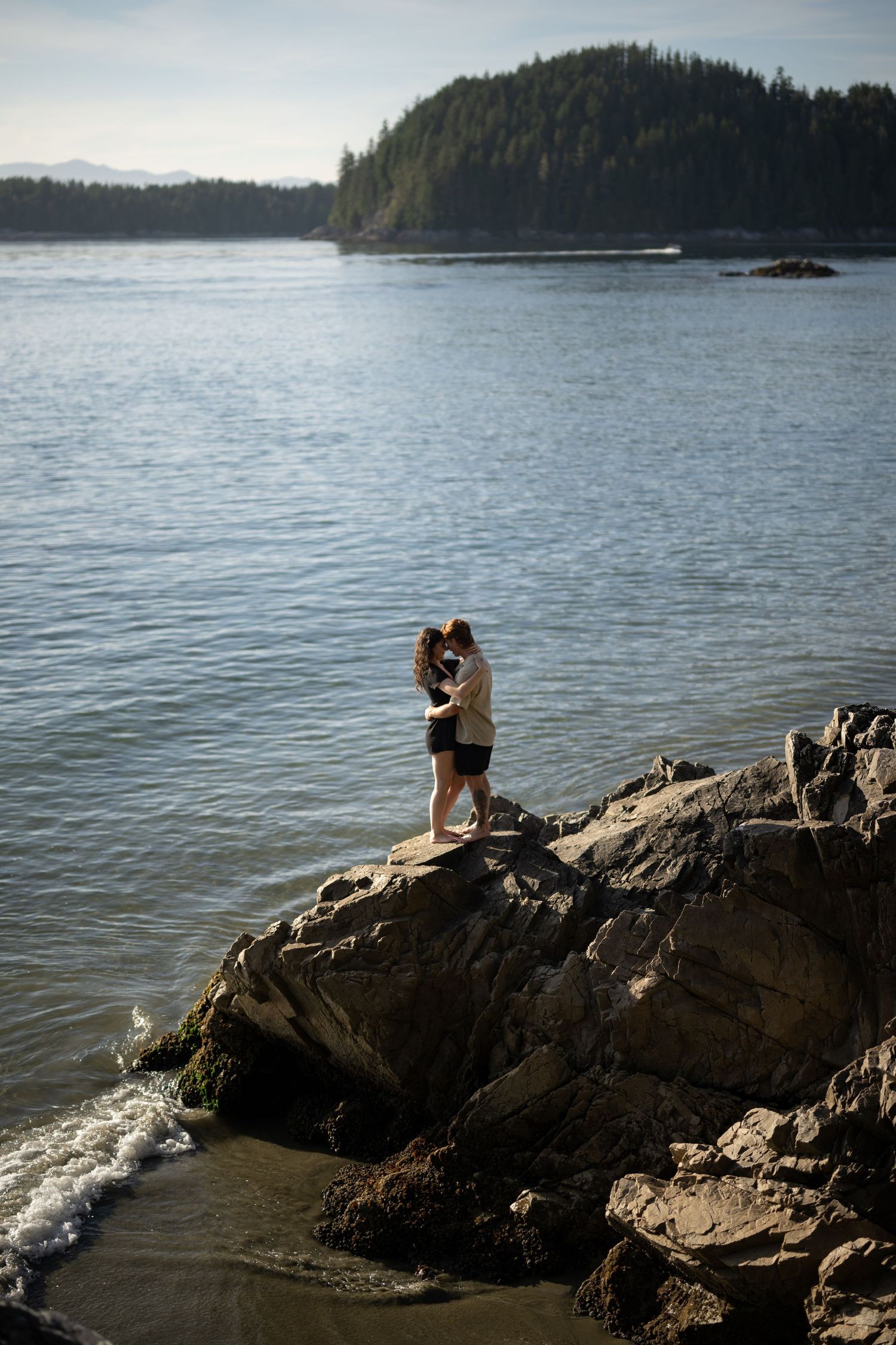 Two figures sit together on rocky outcrop overlooking peaceful ocean bay.