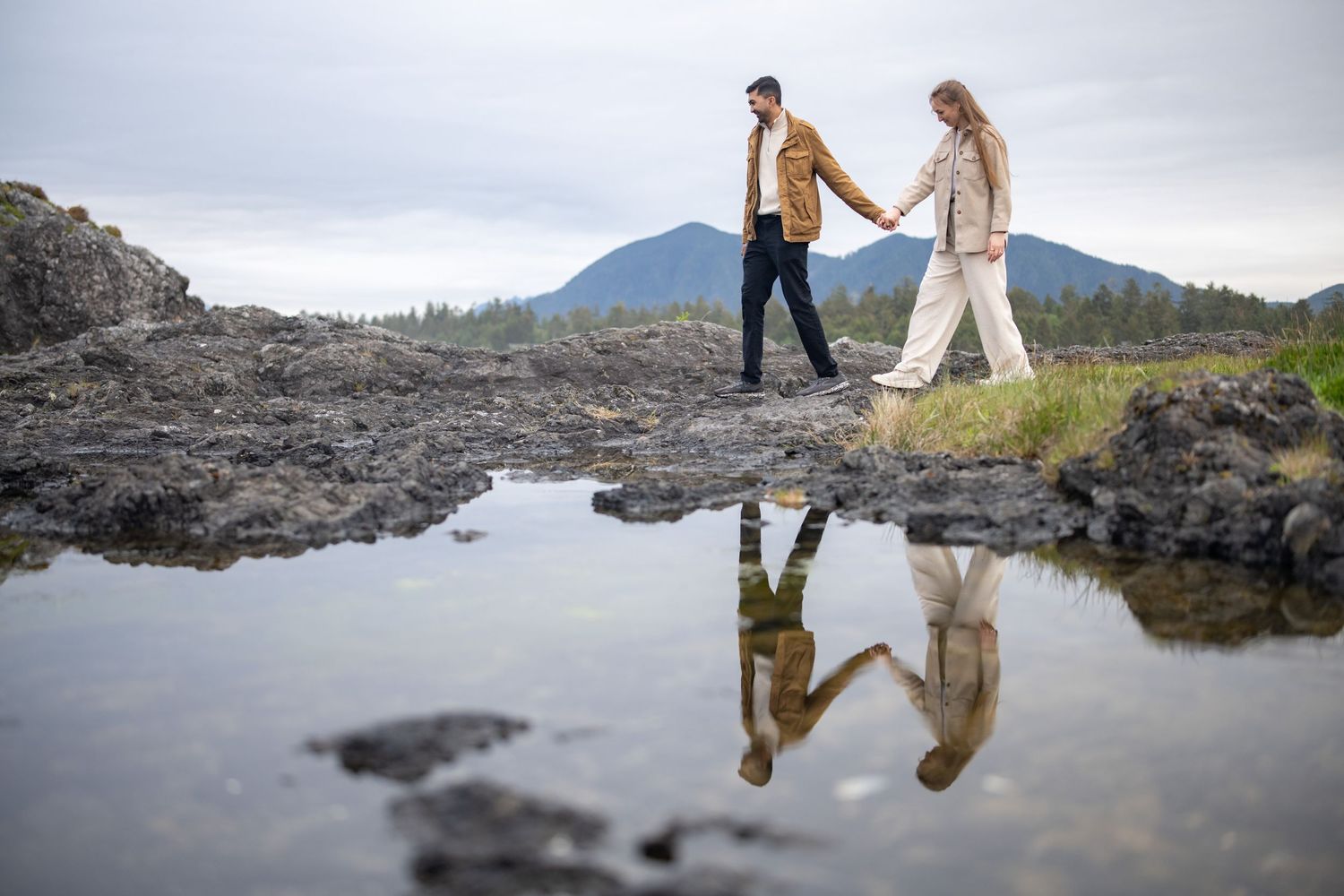 Couple walking along rocky beach with mountain reflections in tide pools.