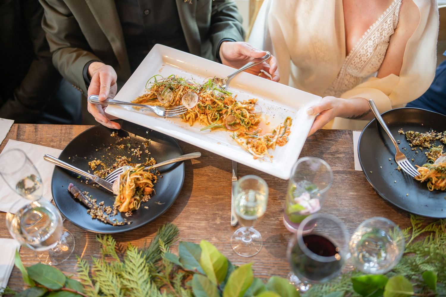 Shared plate of gourmet food being passed around dining table with wine glasses.