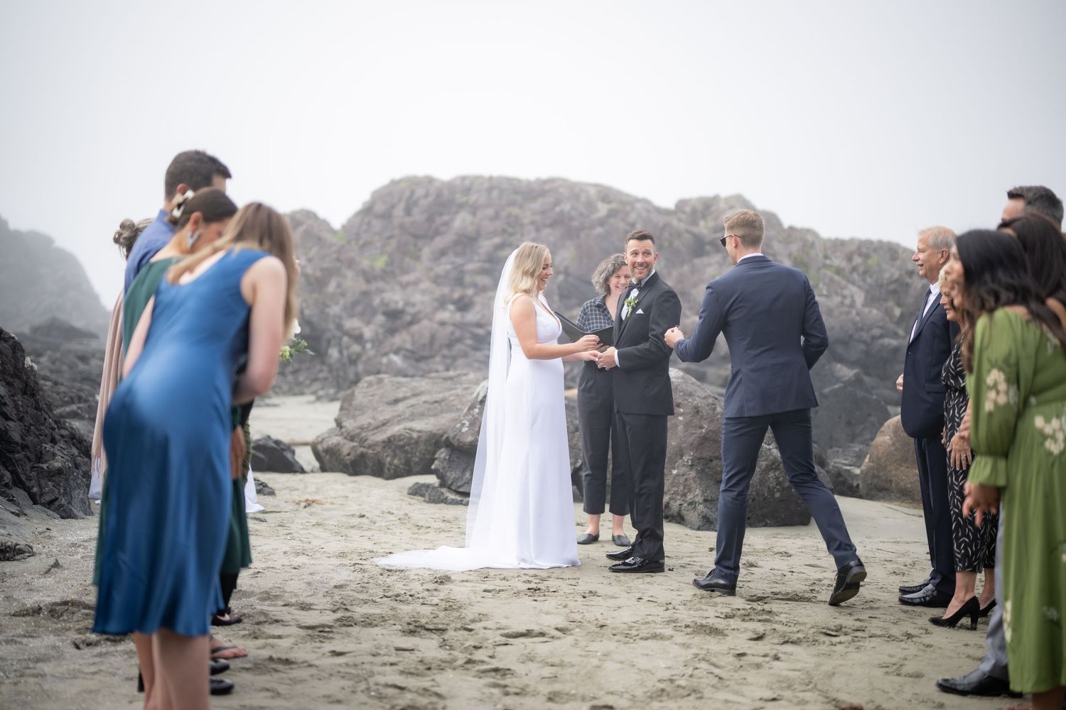 Wedding party and guests celebrate on a misty beach surrounded by dramatic rock formations.