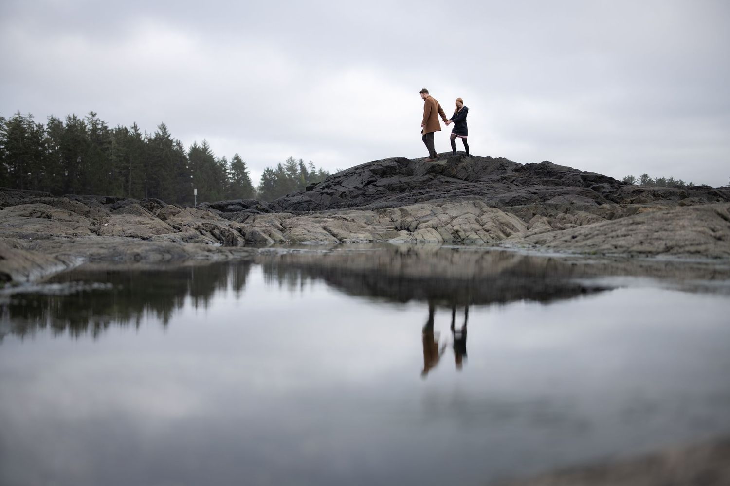 Two silhouettes stand on rocky outcrop reflected in still water under cloudy skies.