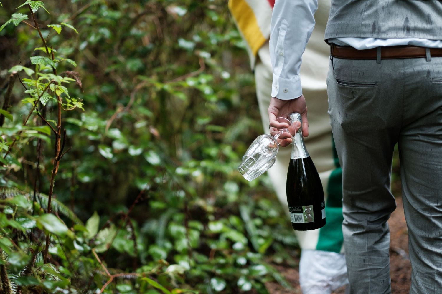 A person in grey pants holds a champagne bottle while walking through greenery.
