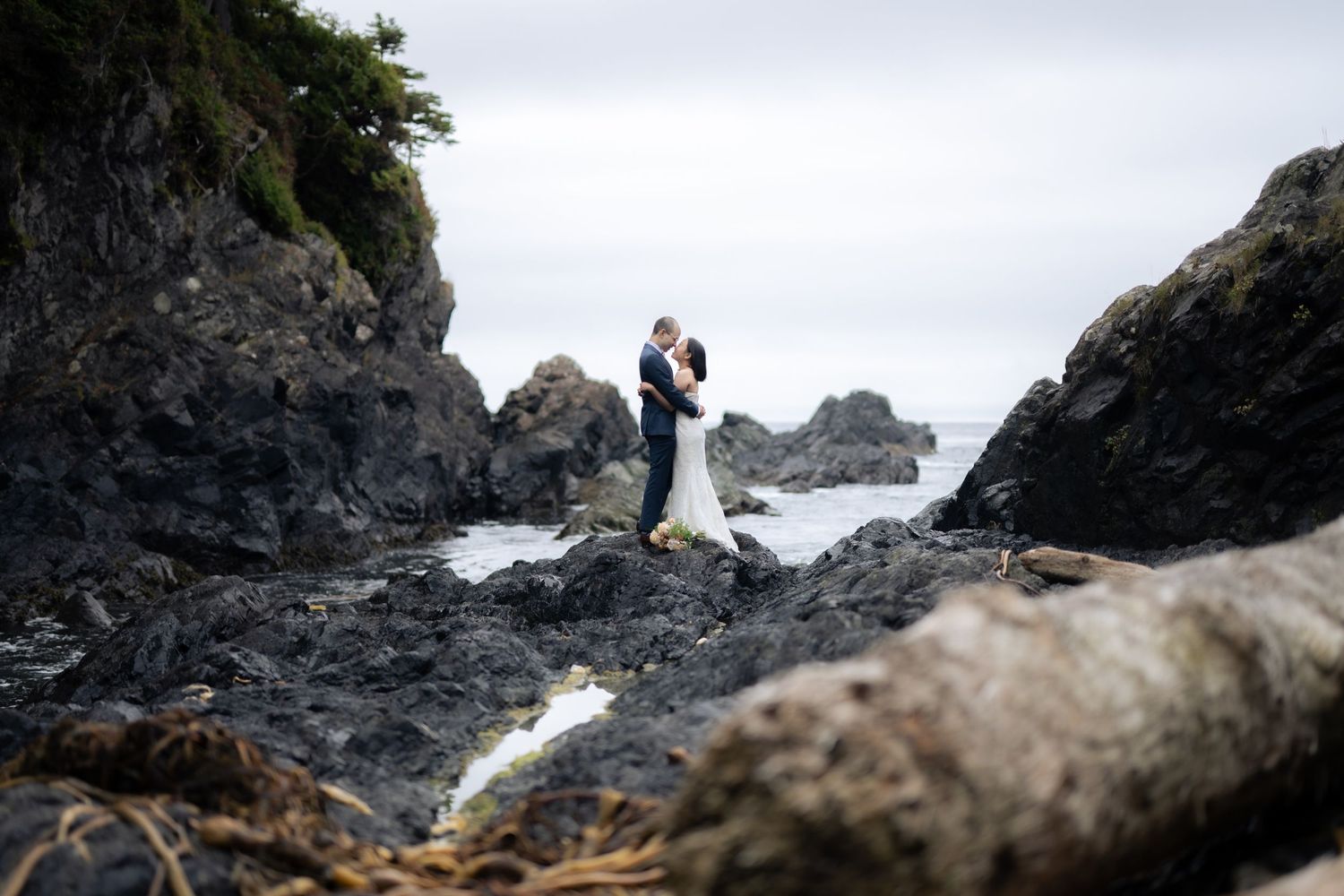 Distant wedding couple silhouetted against rocky coastal landscape and overcast sky.