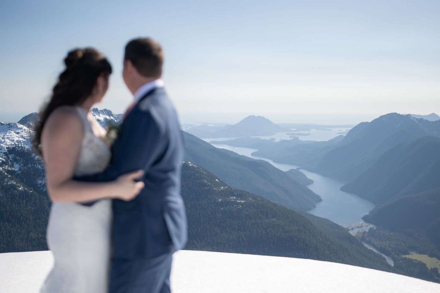 Couple embracing on a mountaintop with dramatic mountain ranges and valleys in the background.