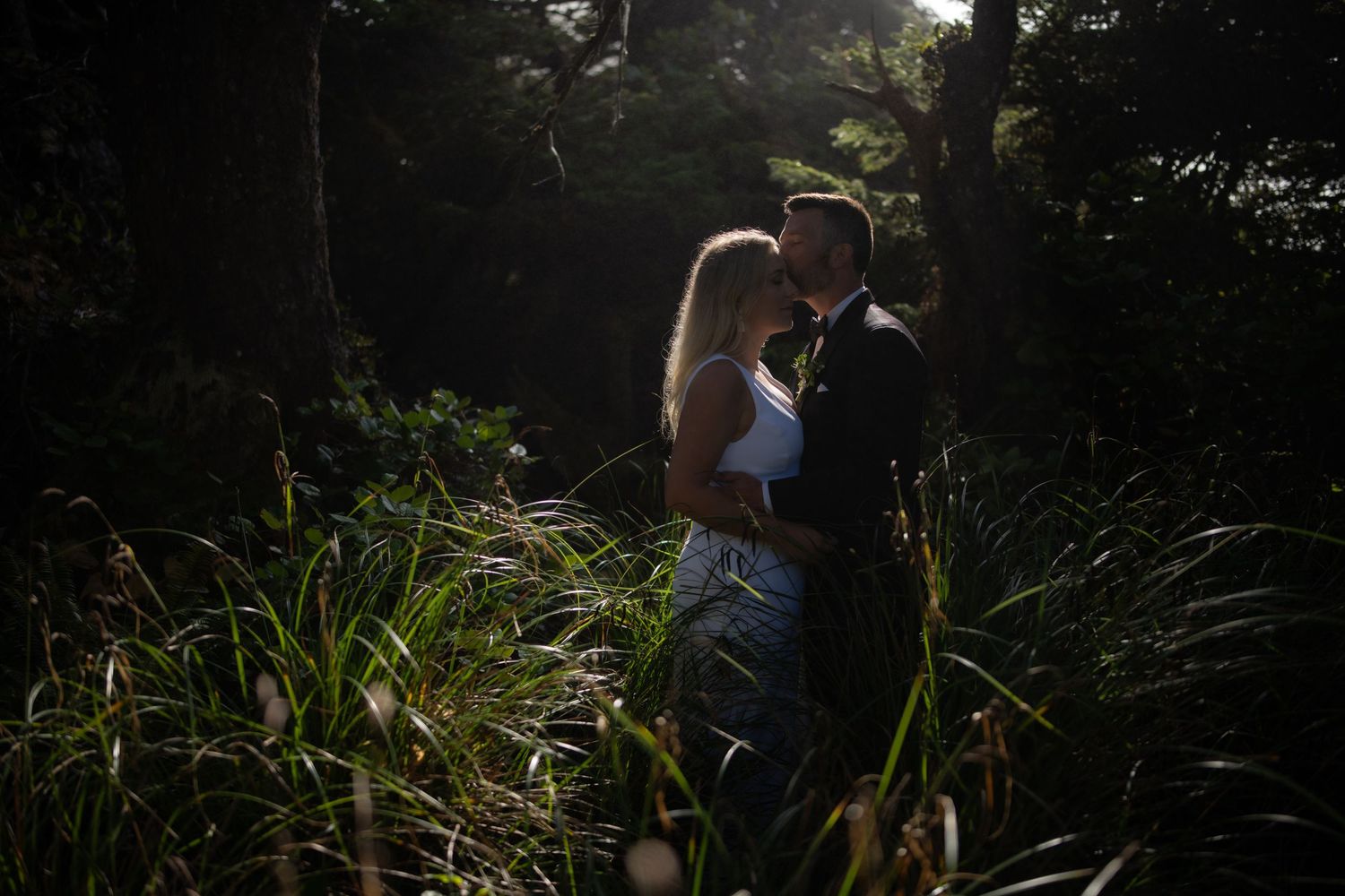 Wedding couple walks through tall grass with sunlight filtering through trees.
