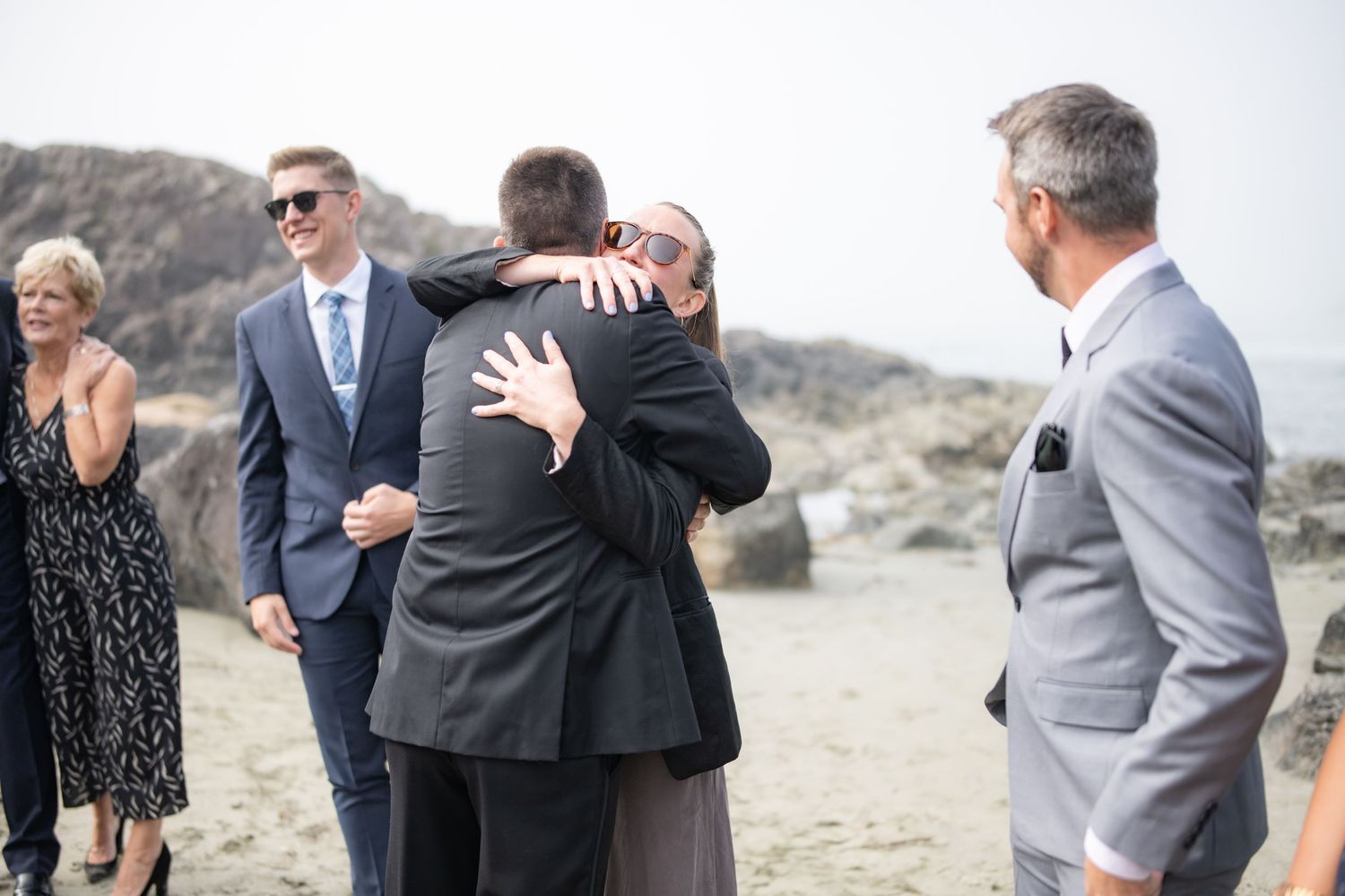 Wedding guests embrace emotionally on a rocky beach with mountains in the background.
