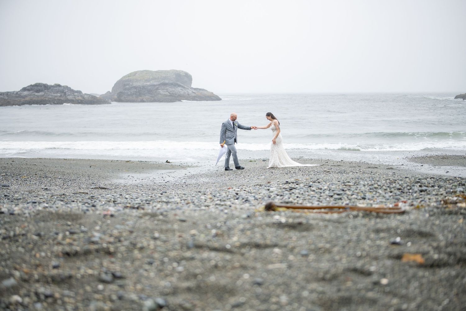 A couple dances together on a misty beach with rocky outcrops in the background.