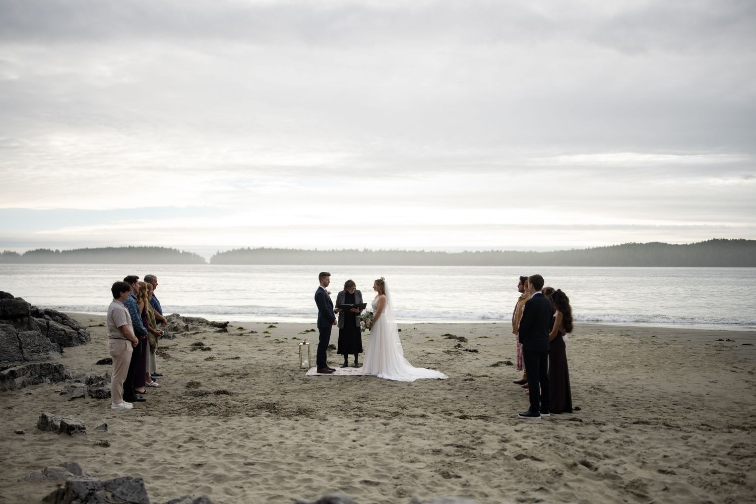 A panoramic view of a beach wedding ceremony with mountains visible across the water.