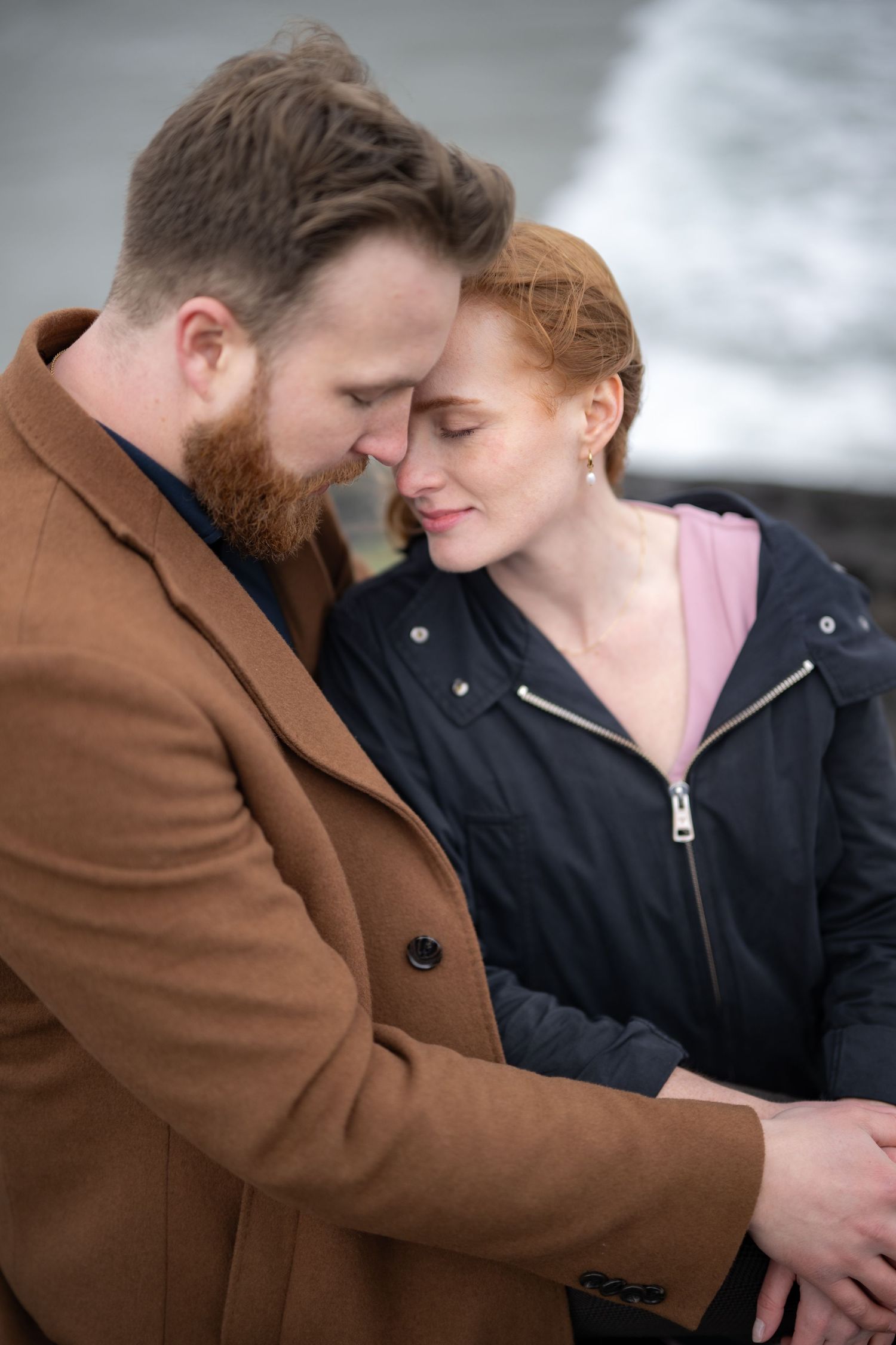 A couple shares an intimate moment wearing brown and black coats against a misty backdrop.