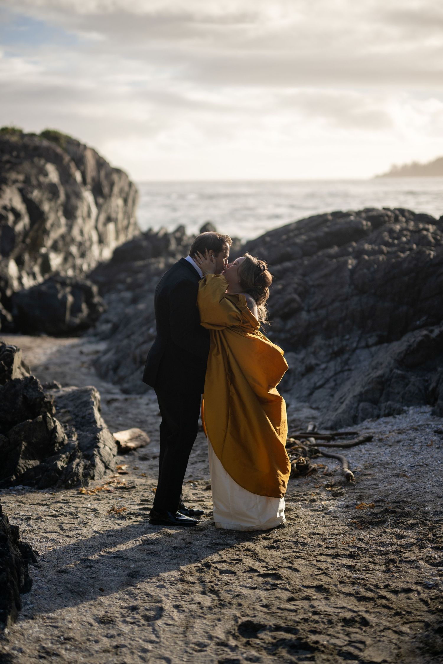 A couple sits together on coastal rocks overlooking the ocean at dusk.