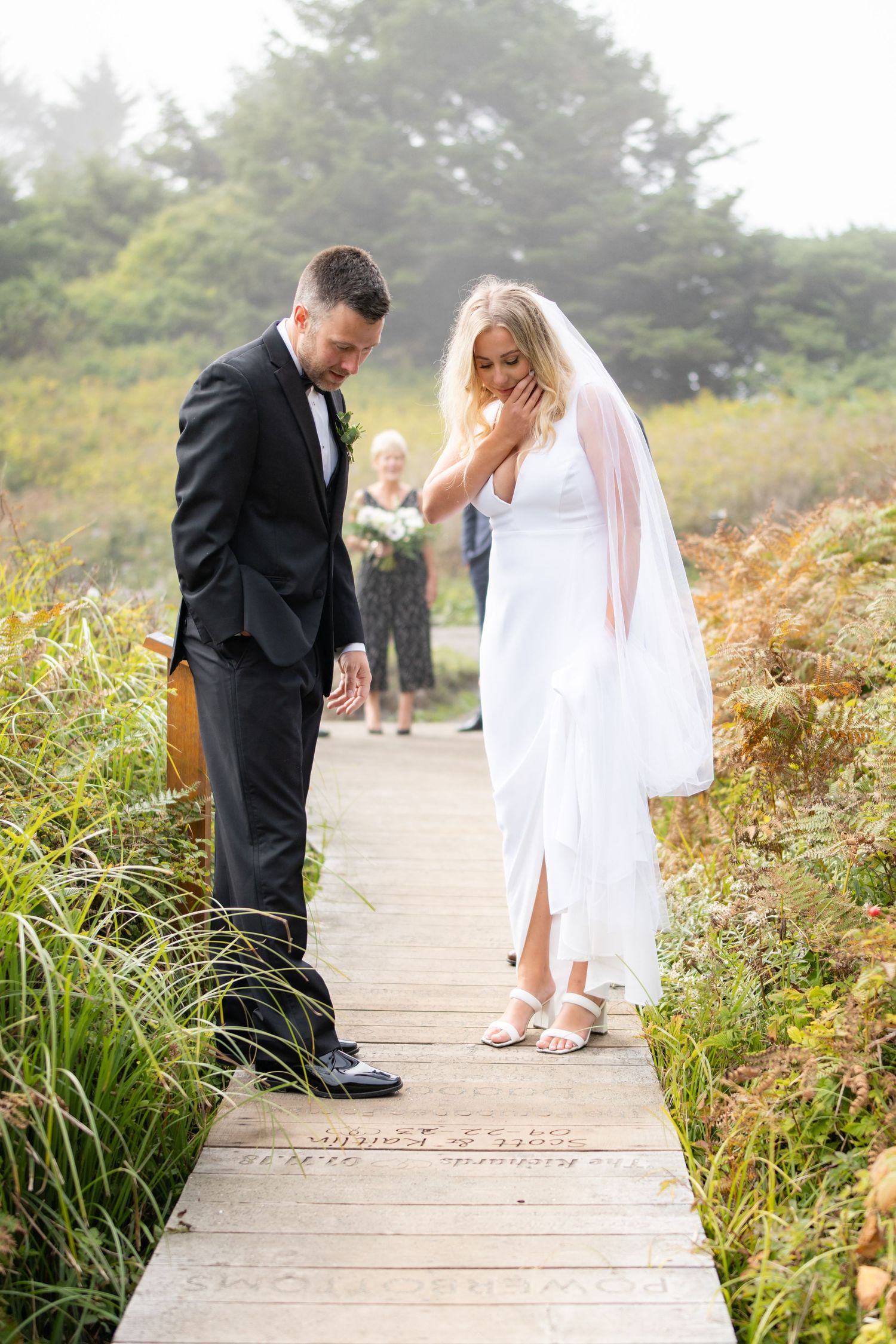 Couple walking along wooden boardwalk surrounded by coastal vegetation.