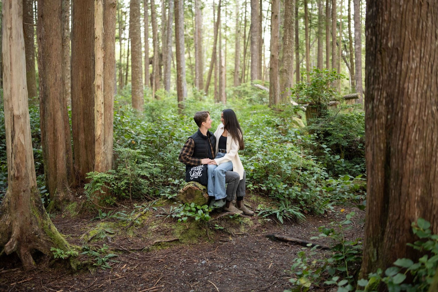 Wide forest landscape photo showing a couple among tall evergreen trees on a dirt path.