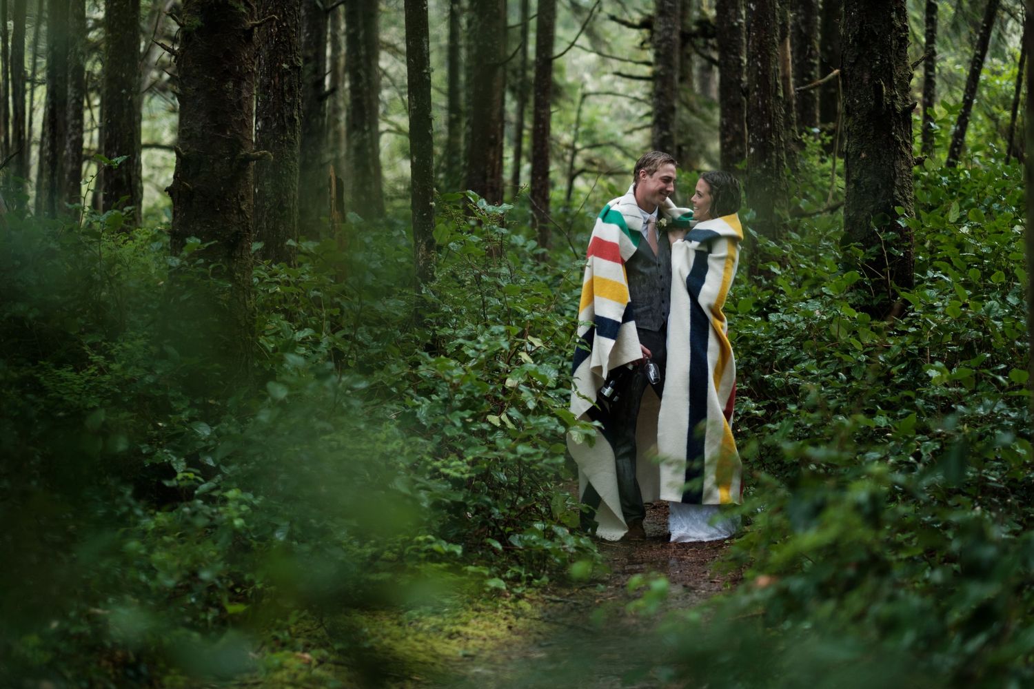 A couple stands together on a forest path wrapped in a colorful striped blanket.