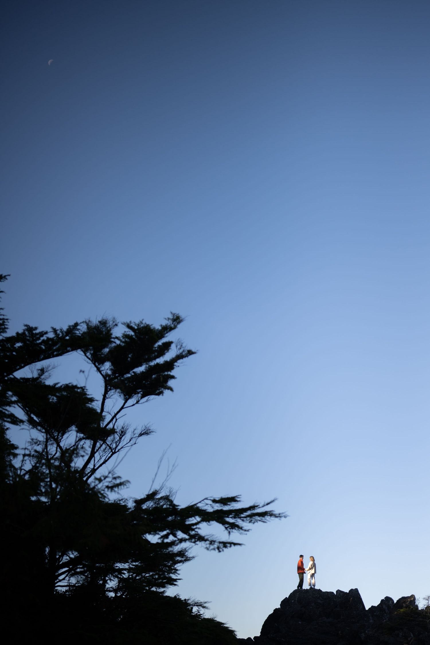 Two silhouettes stand atop a cliff against a deep blue sky at dusk.