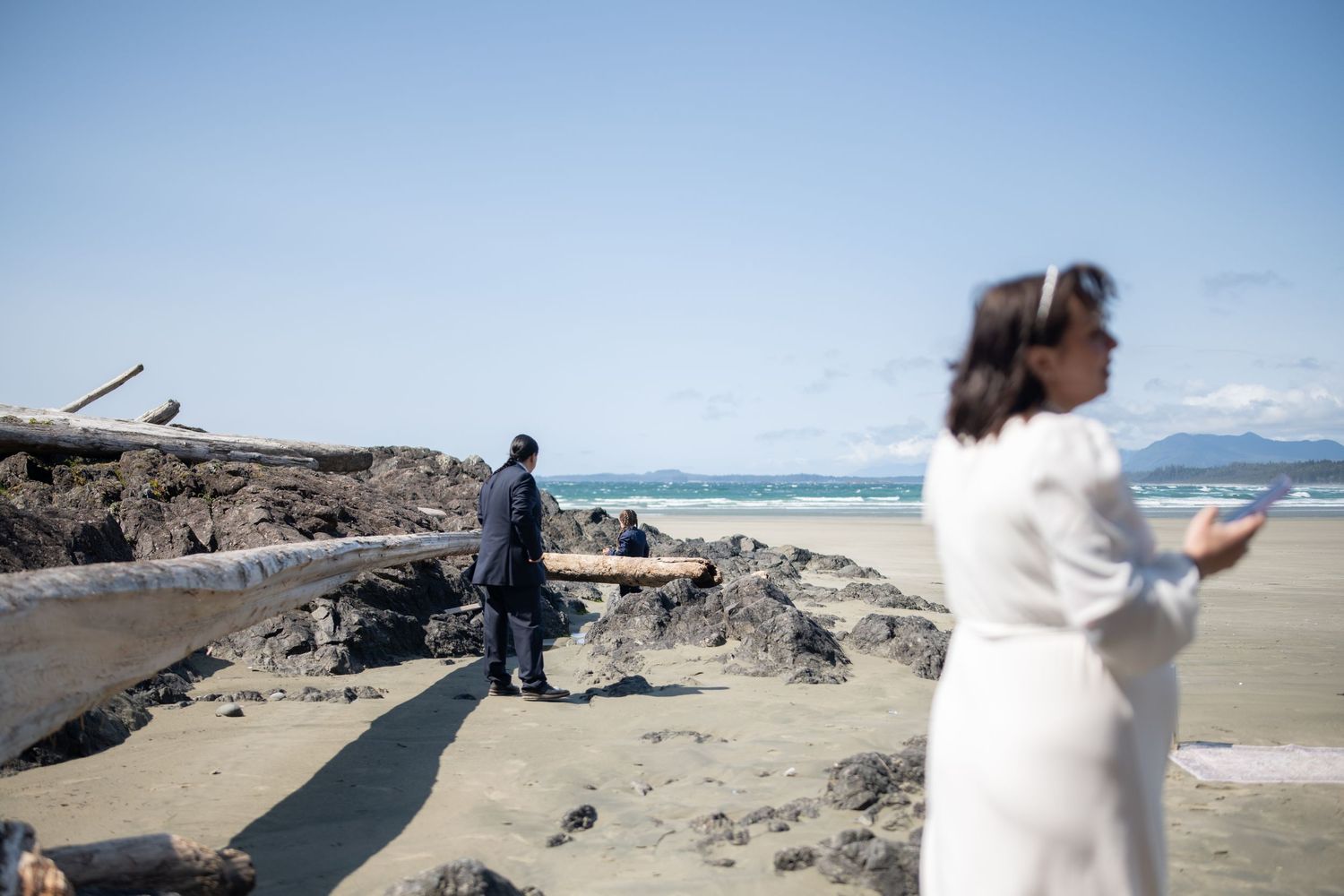 Wedding ceremony participants gather on a scenic beach with mountains in the background.