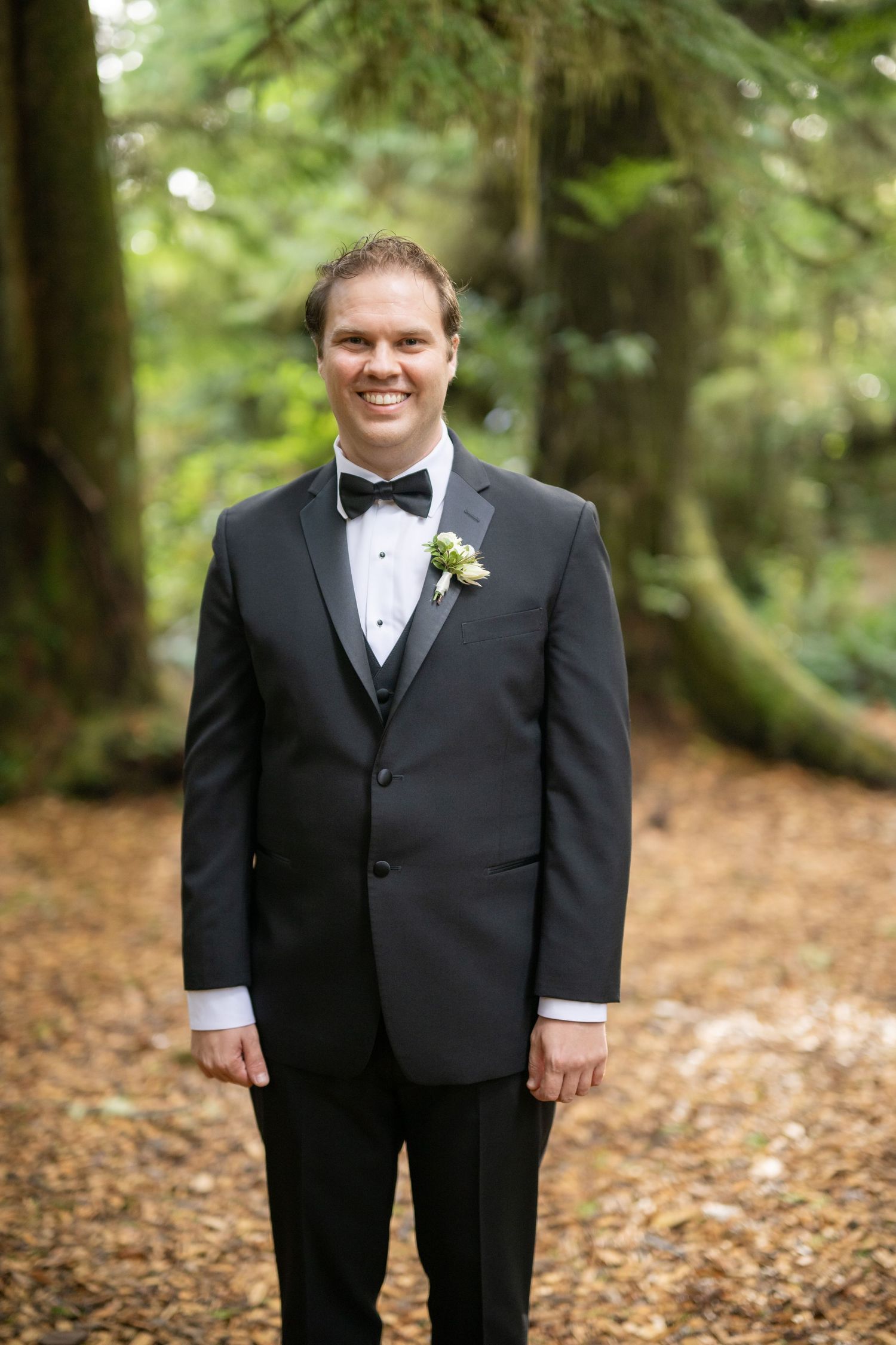 A formal portrait taken on a leaf-covered forest path with classic black tuxedo attire.