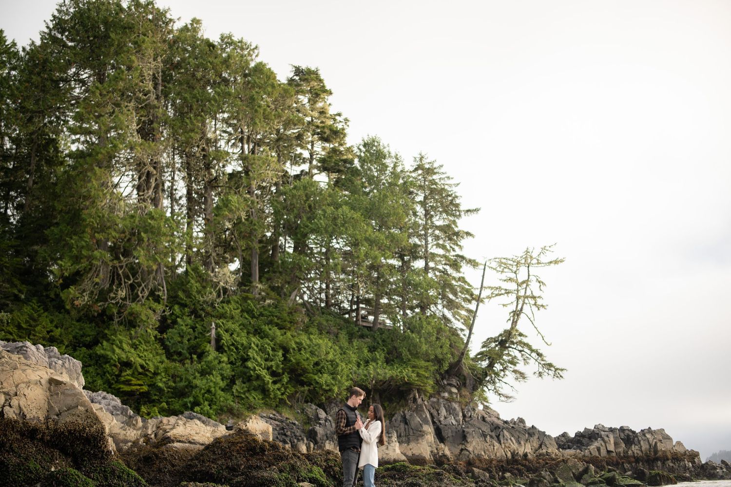 Couple standing together on rocky coastal cliff surrounded by evergreen trees.