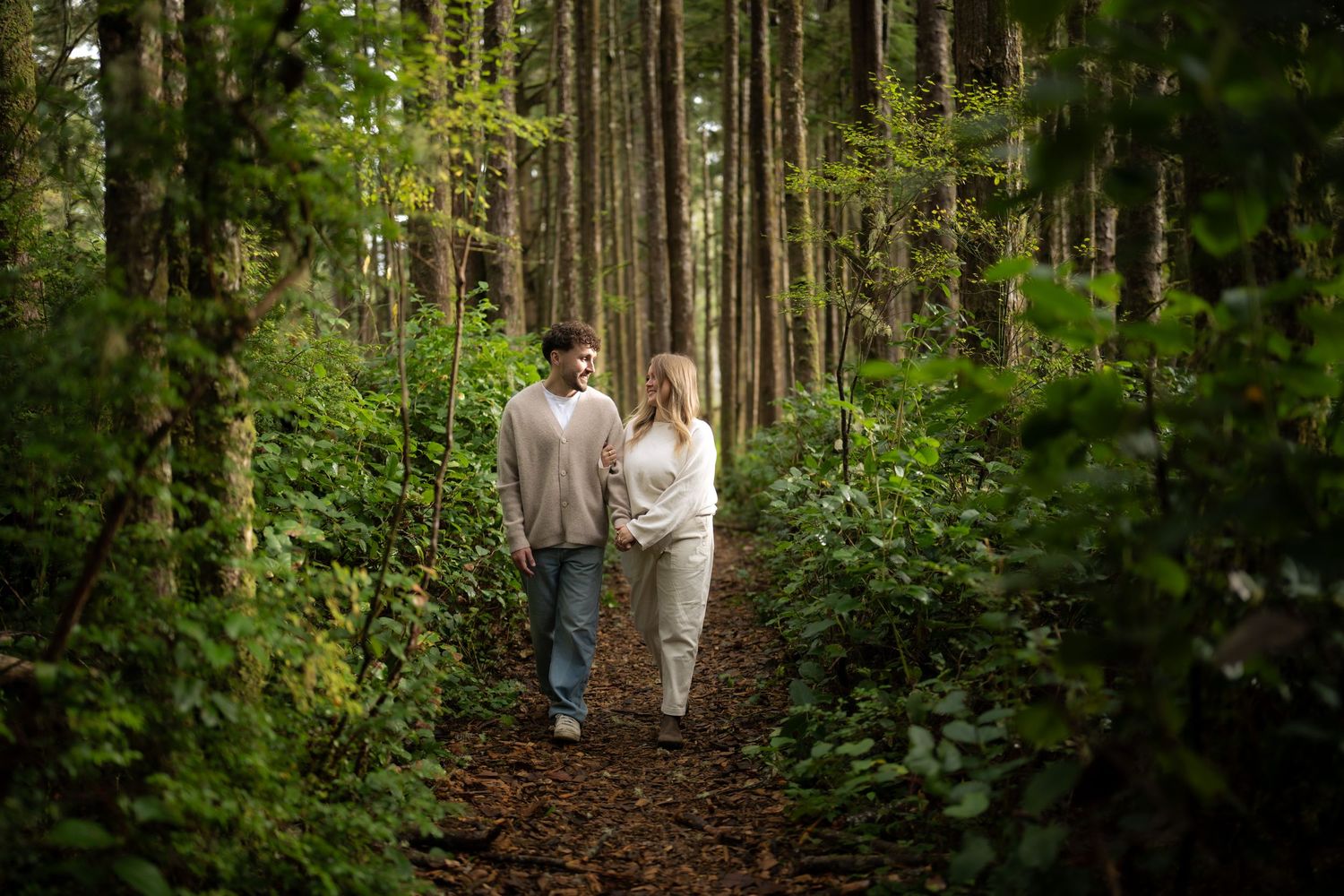 A romantic forest walk along a dirt trail surrounded by towering evergreens and dense foliage.