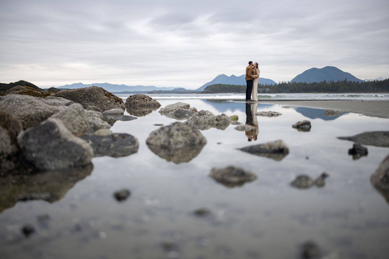 Scenic beach landscape with rocks and mountains reflected in shallow water.