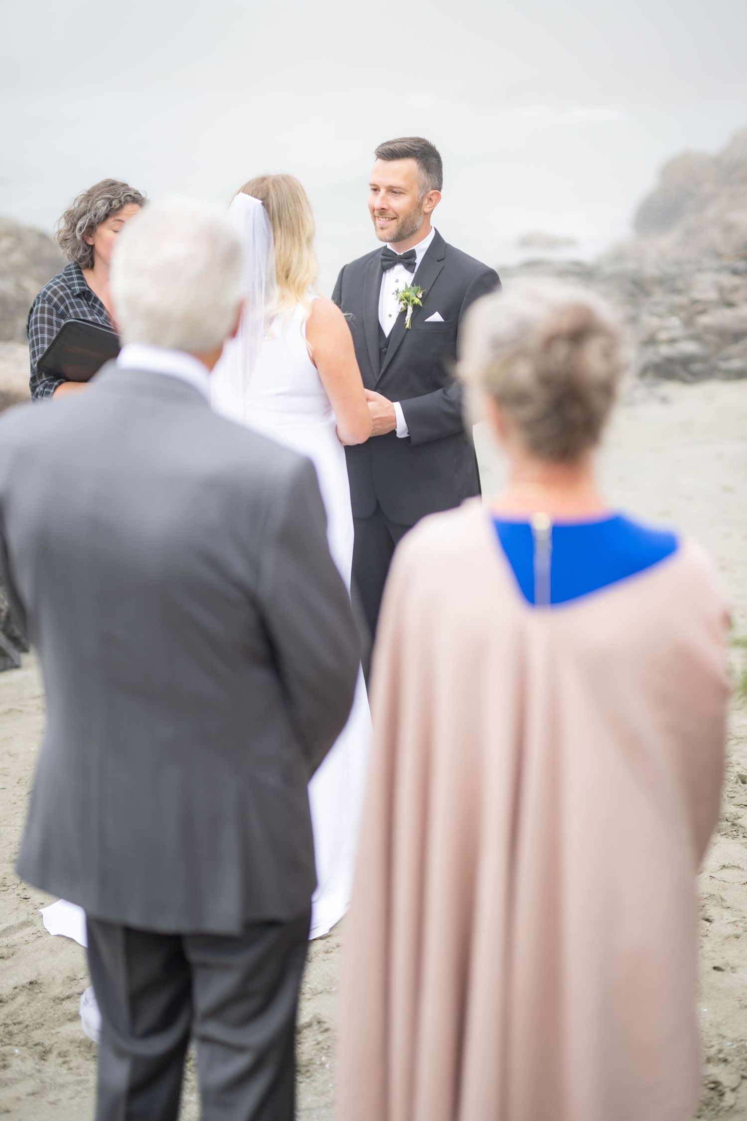 Wedding guests gather on a rocky beach during an outdoor ceremony as fog rolls in.