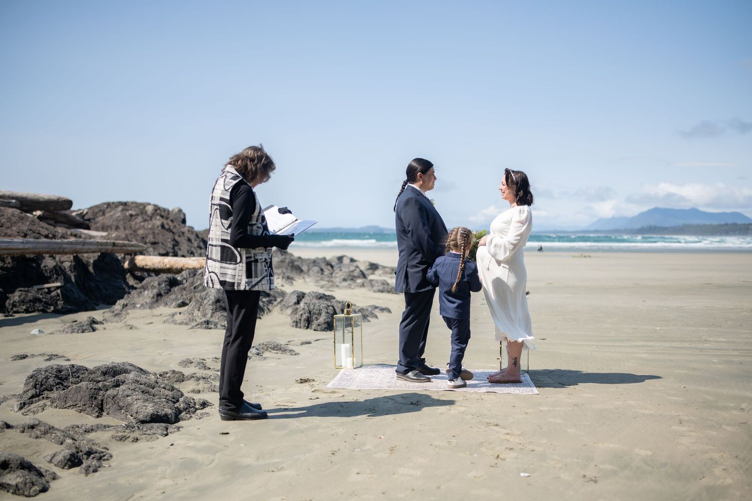 Wedding guests gather on a scenic beach with rocky outcrops and mountains in the distance.