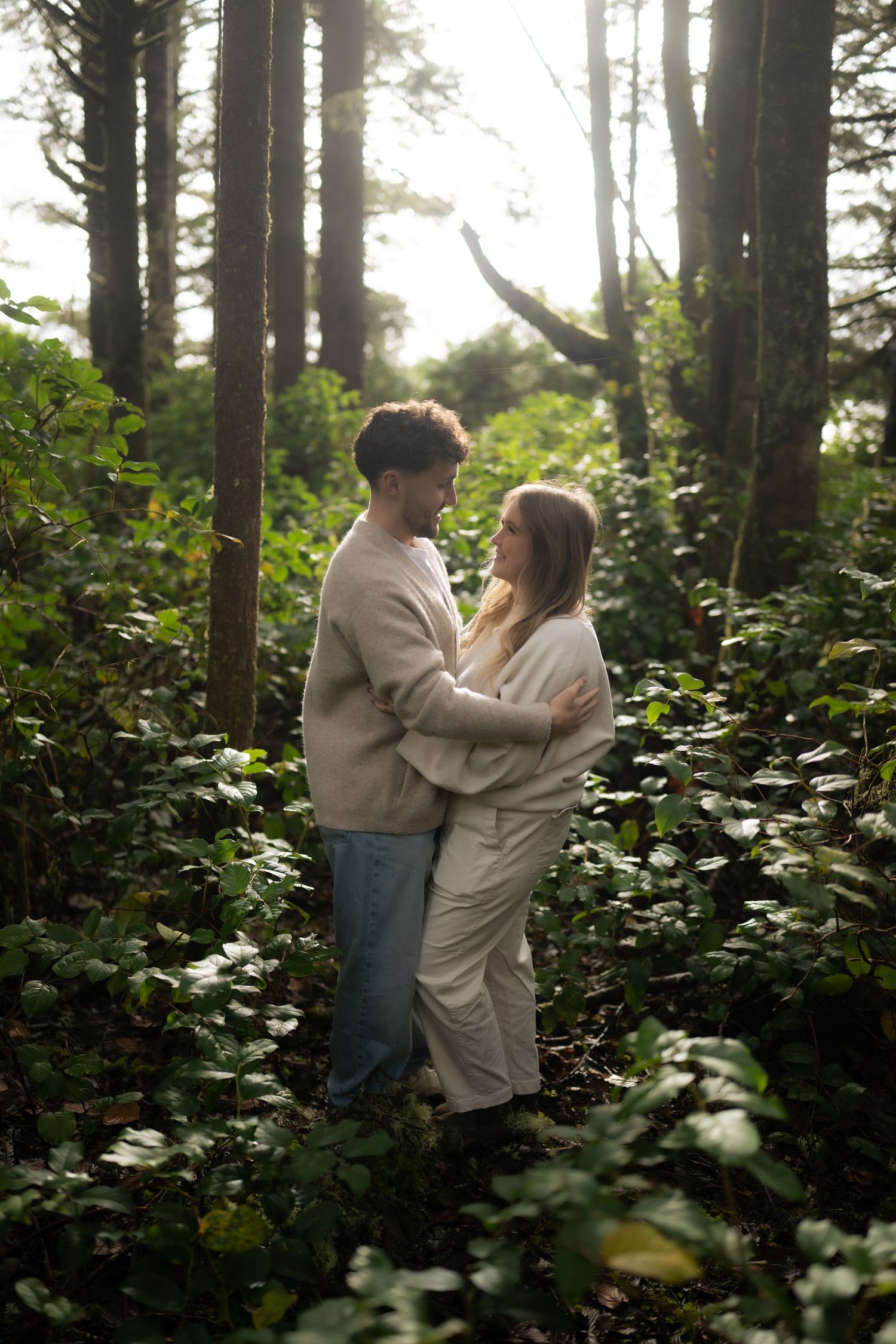 Sunlit forest scene with two people sharing tender moments among evergreen trees and lush foliage.