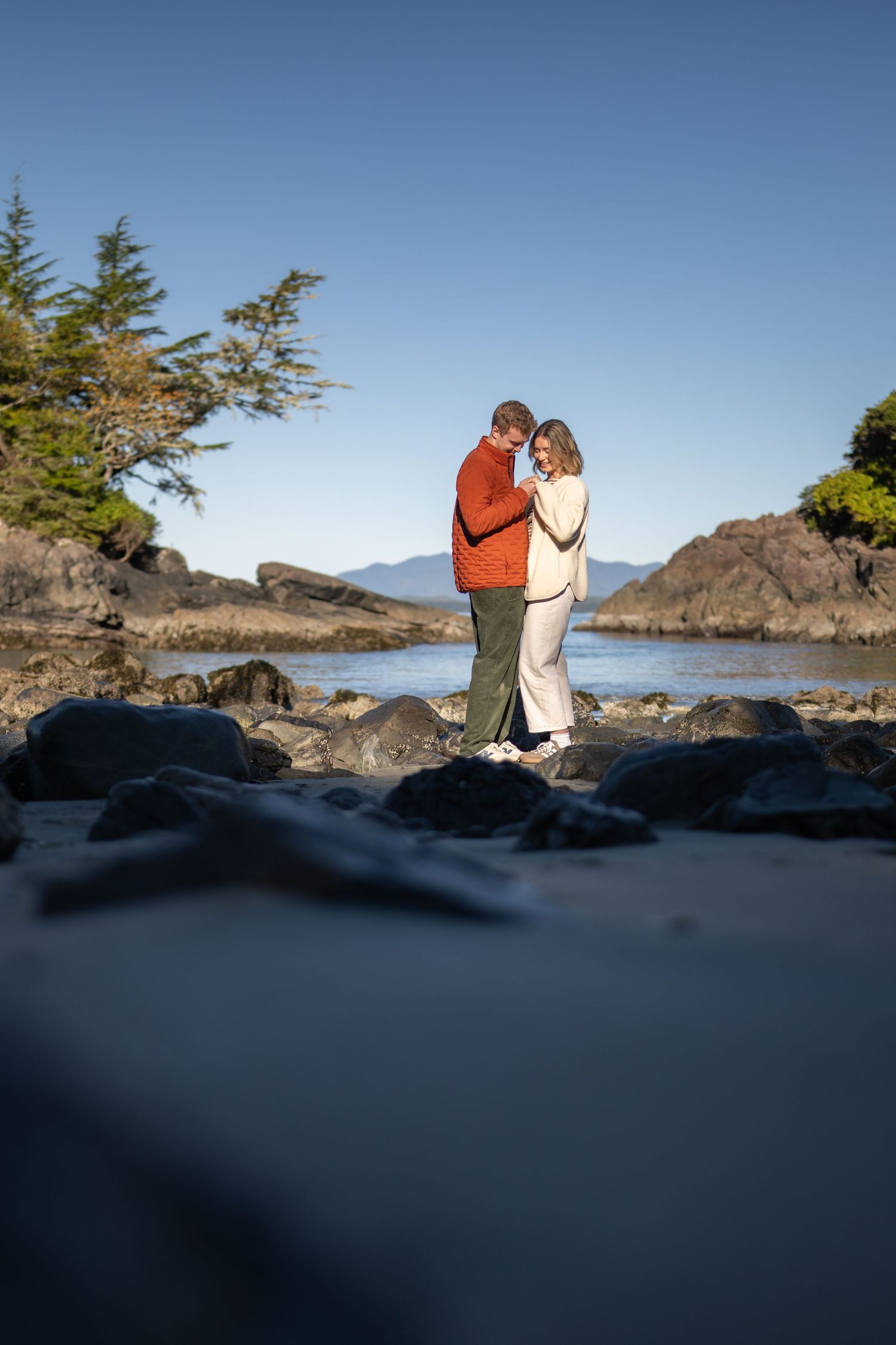 A couple embraces on rocky coastal cliffs against a bright blue sky.