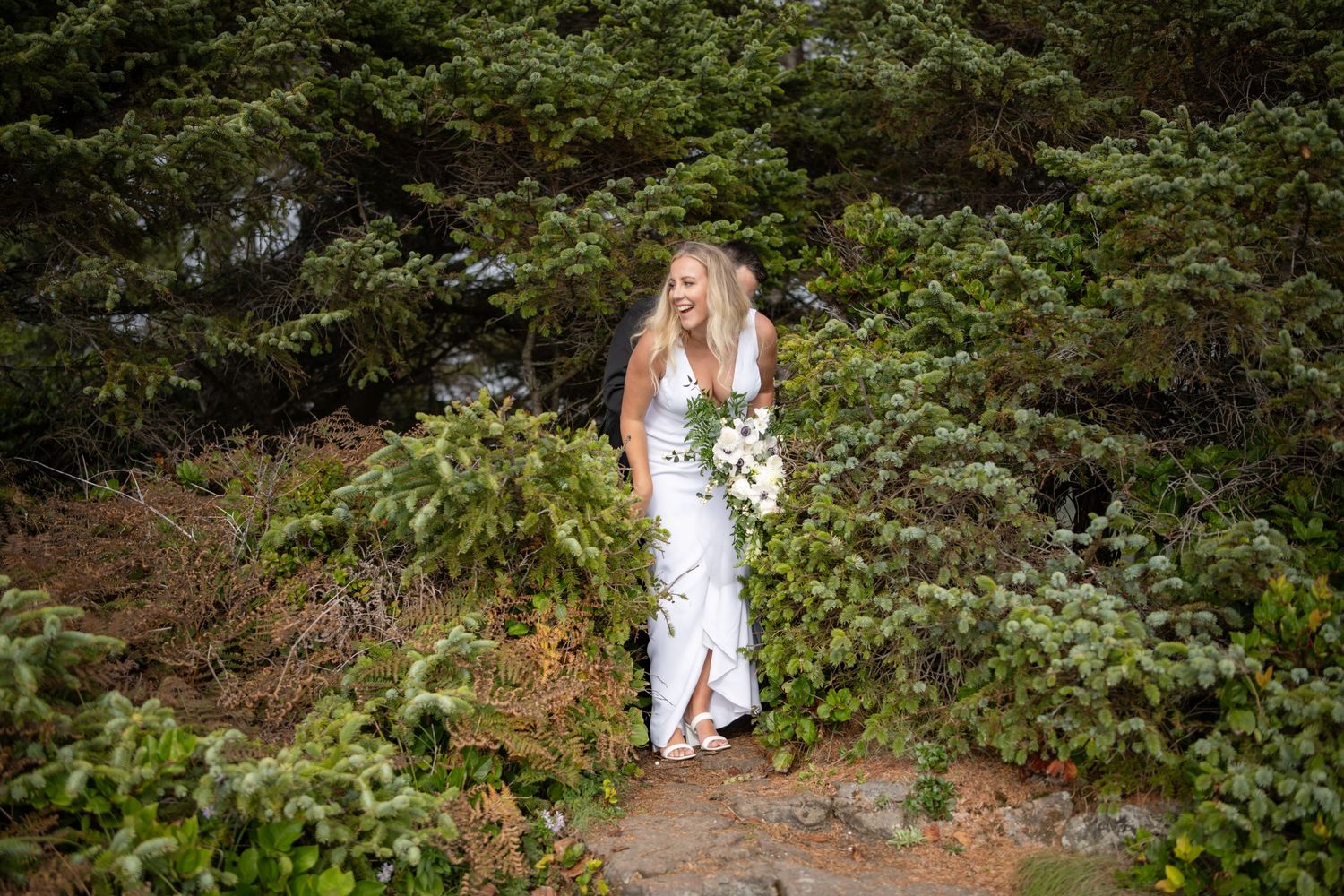 A person in a white dress walking along a rugged mountain path surrounded by greenery.