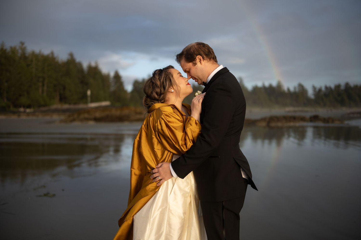 A romantic moment captured on a moody beach with forest backdrop and rainbow overhead.