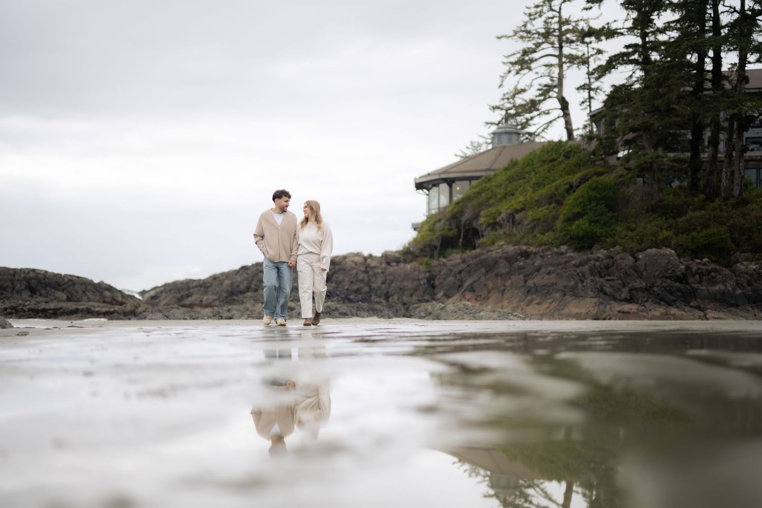A couple in cozy sweaters walking along a misty Pacific Northwest beach with their reflection in wet sand.