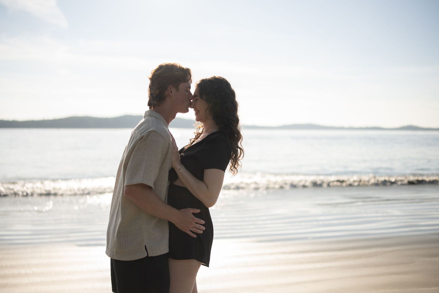 Two people share an intimate moment on a sunny beach with waves in the background.