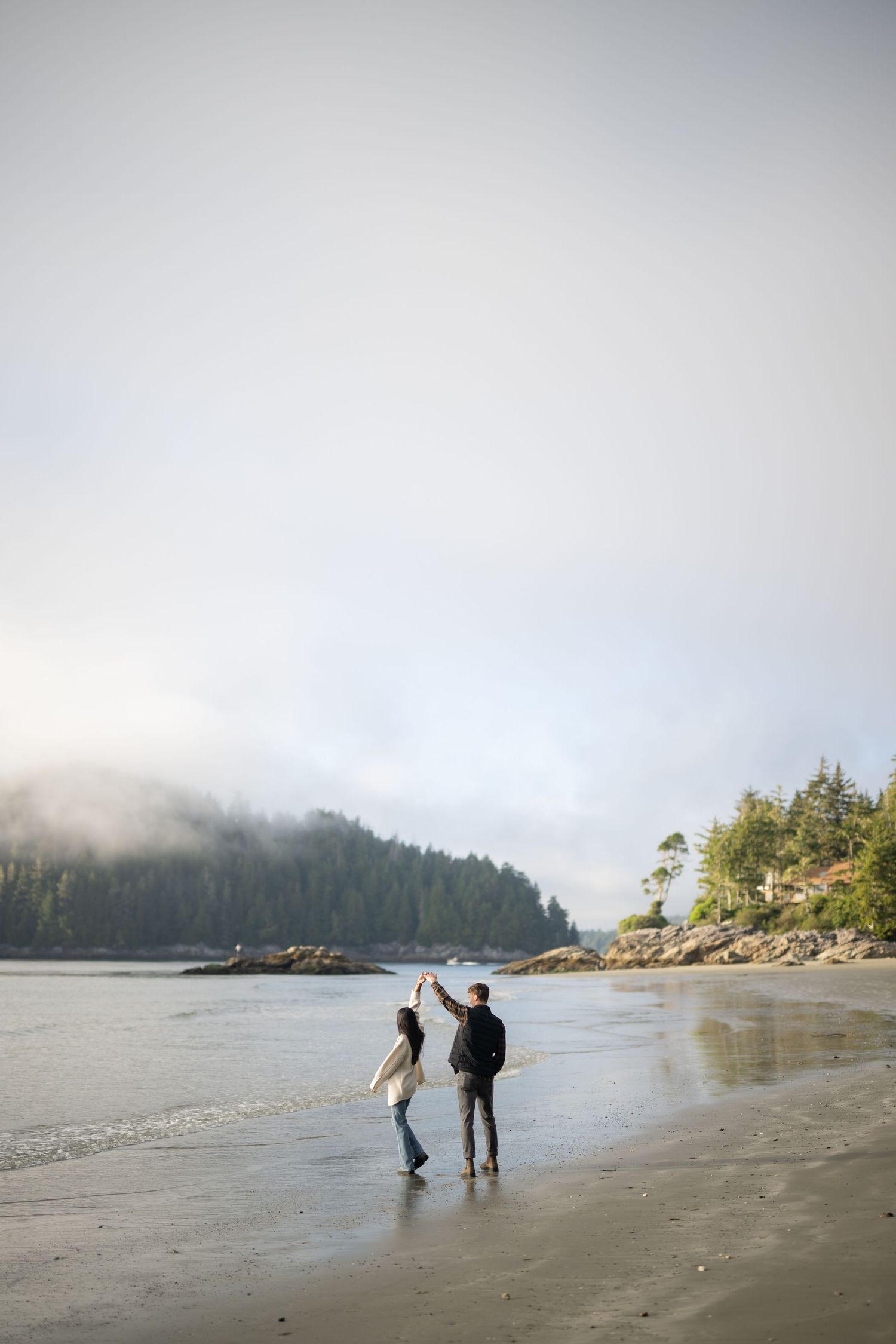 Two silhouettes walking on misty beach with evergreen forest backdrop.