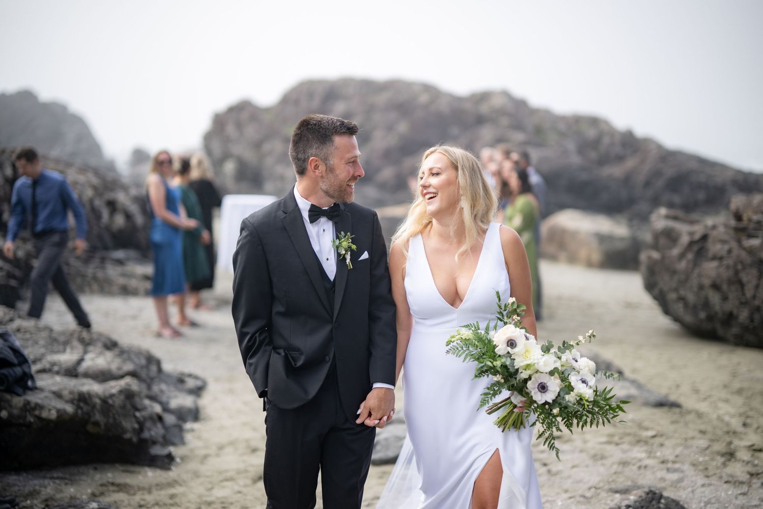 Elegant beach wedding couple poses together against dramatic coastal rocks.