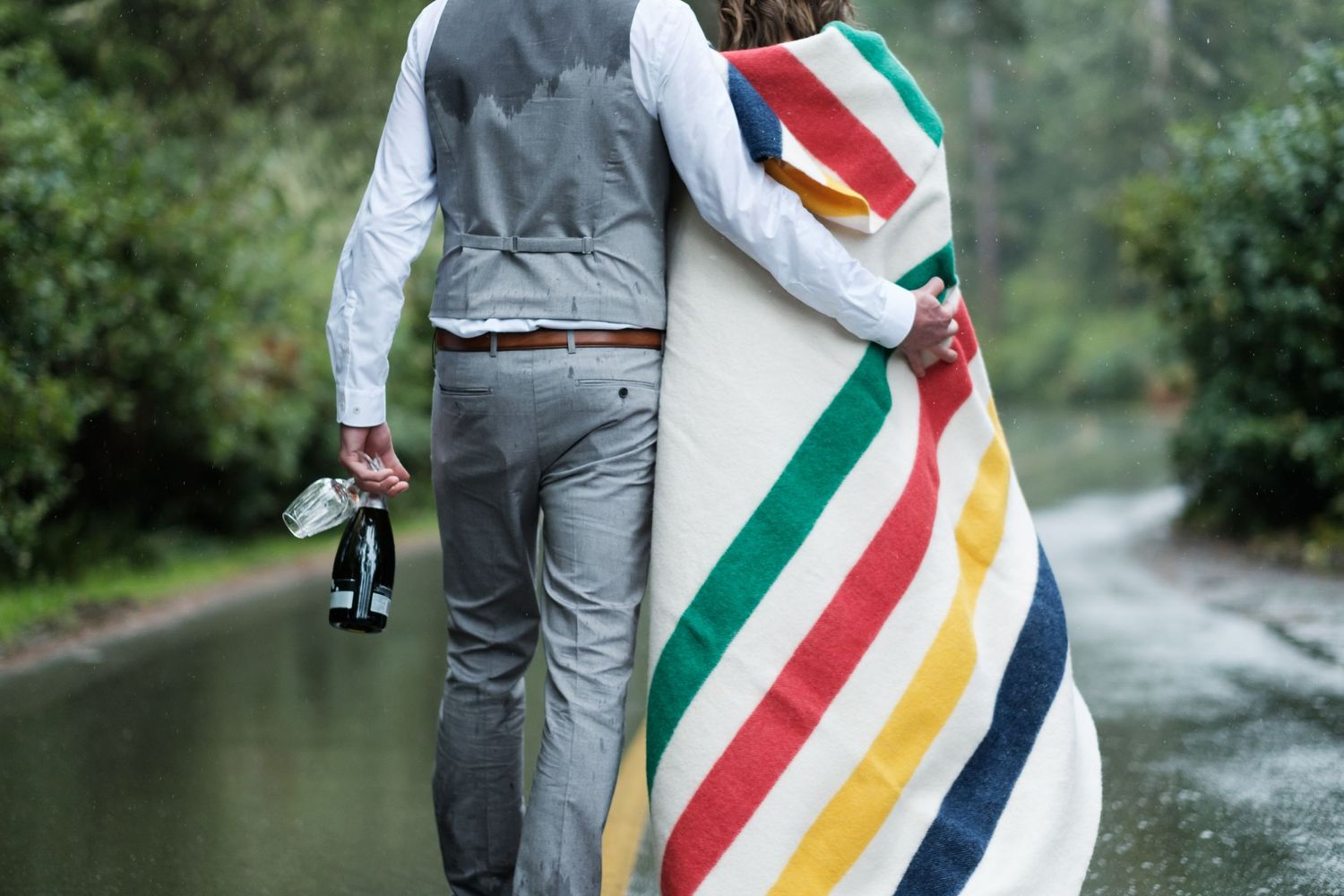 A couple holding photography equipment walks in the rain with a striped blanket.
