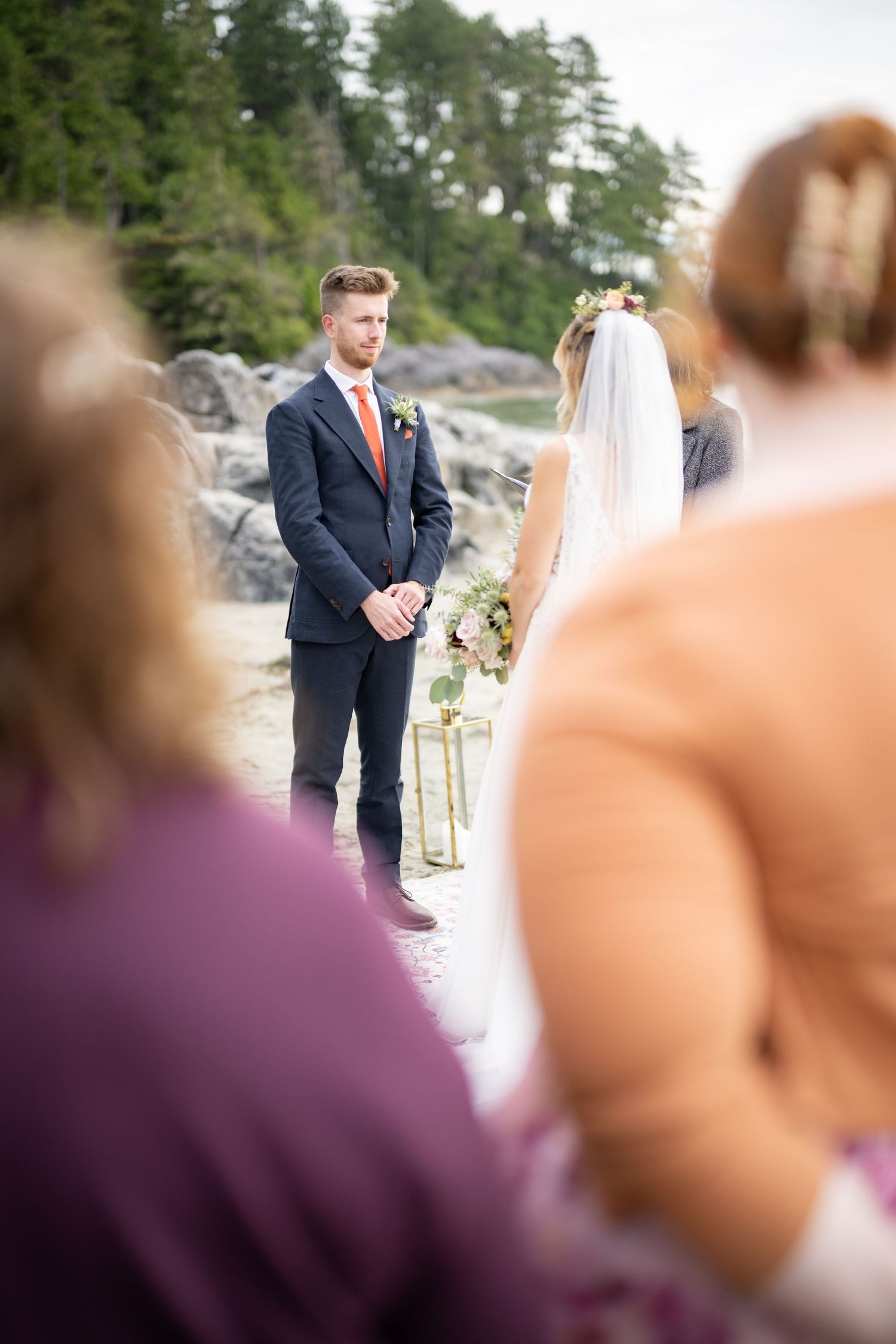A wedding ceremony unfolds on a beach with evergreen trees and rocky shoreline in the background.