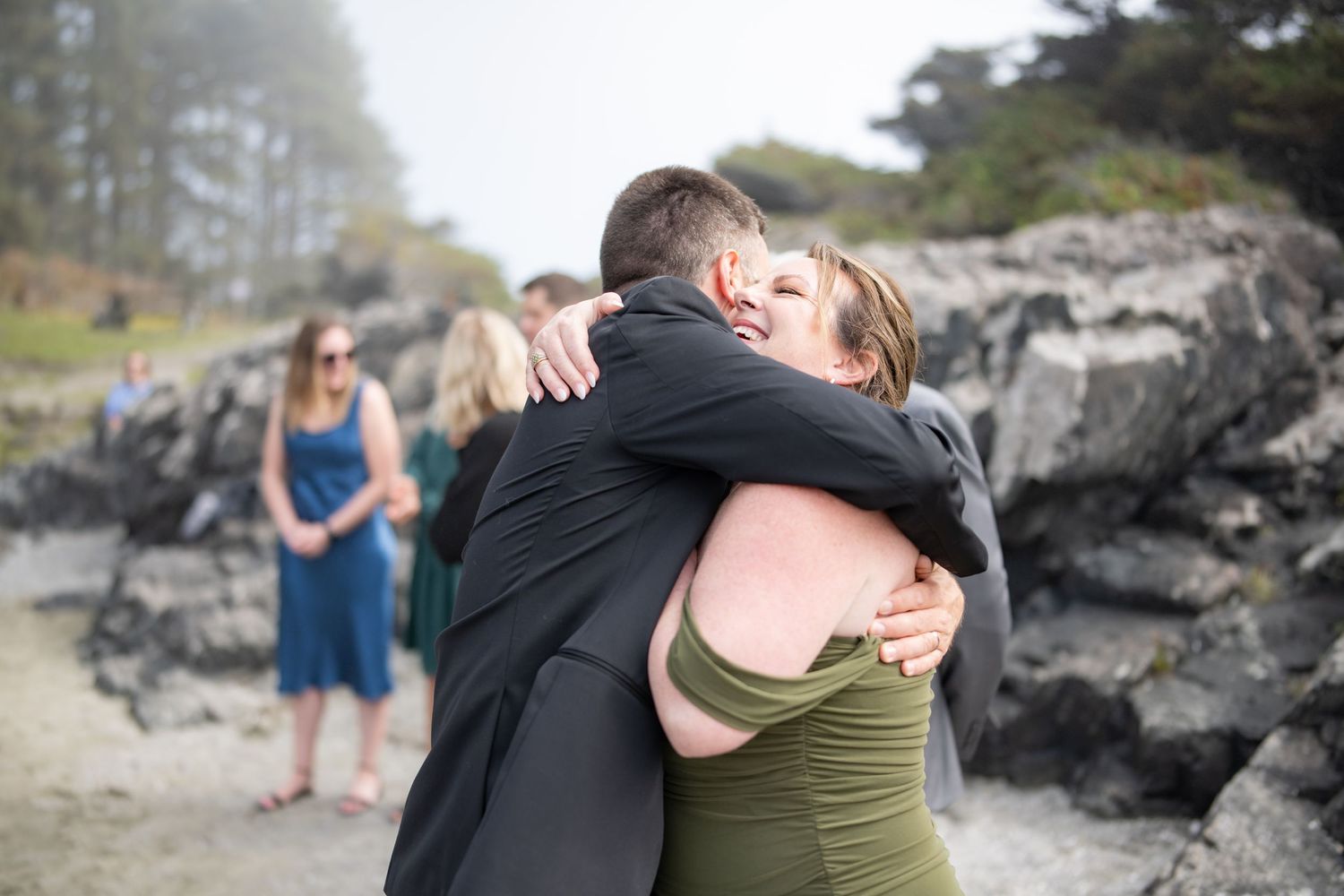 Emotional embrace between wedding attendees on a misty beach with rocky cliffs.